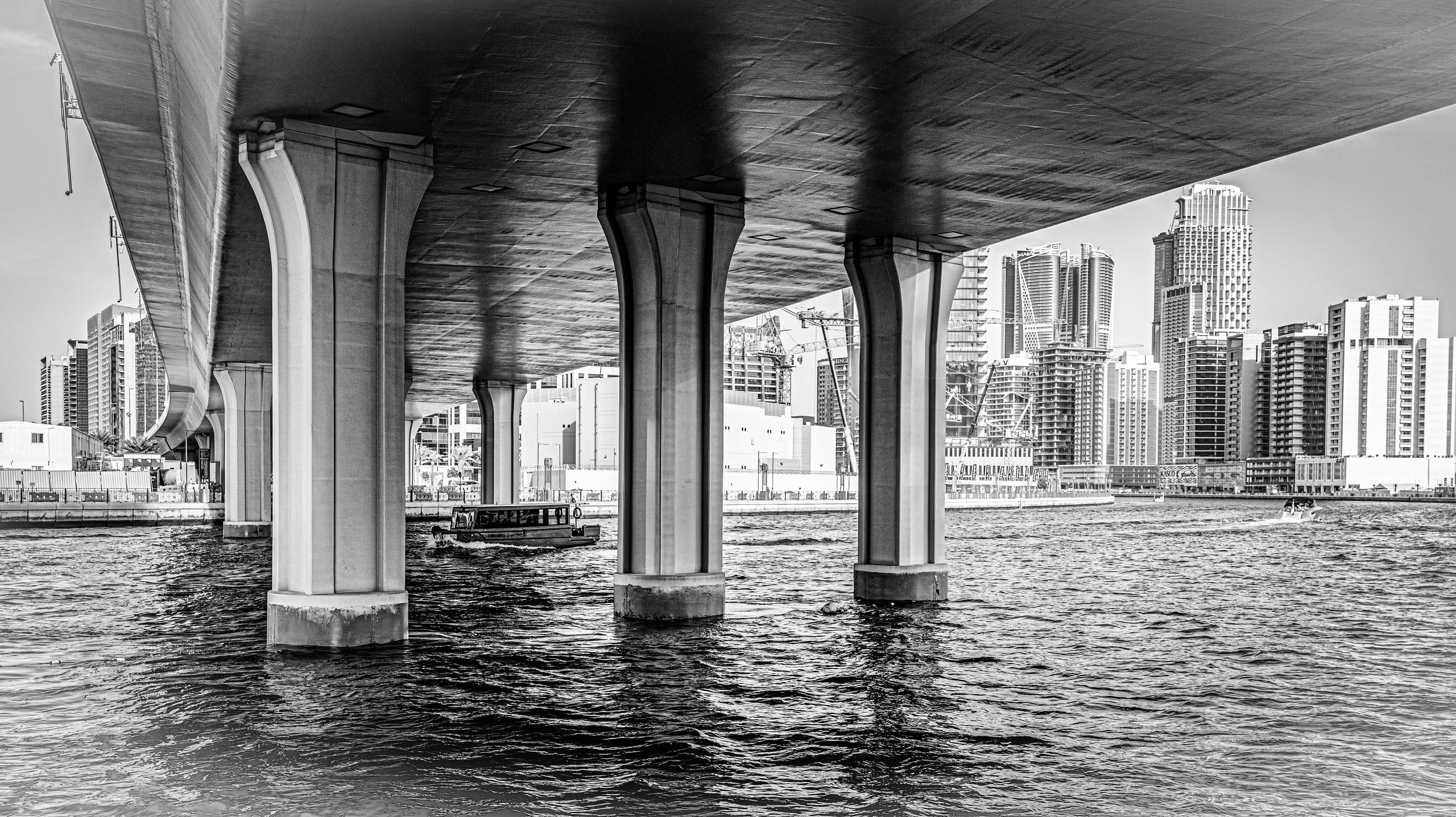 Black-and-white architectural view looking underneath a large highway bridge supported by massive concrete columns planted in dark water, with a small boat crossing beneath and high-rise buildings visible in the background.