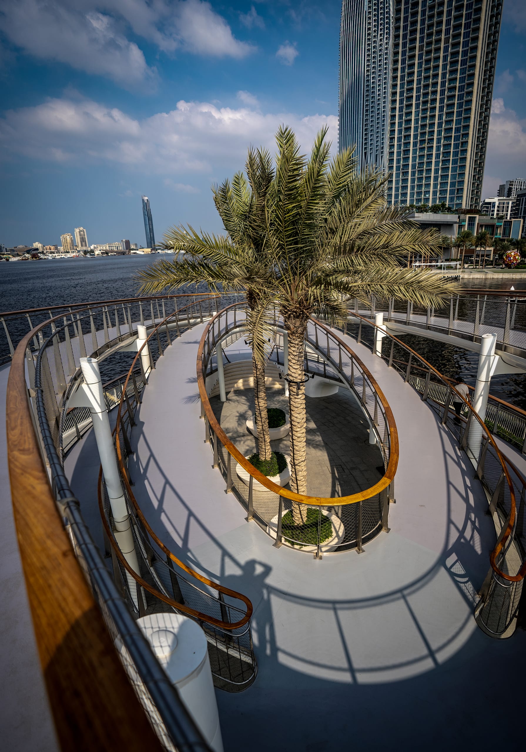High-angle view of an elliptical pedestrian overpass with curved wooden railings and two palm trees growing in a central planter, overlooking water and modern skyscrapers in Dubai.