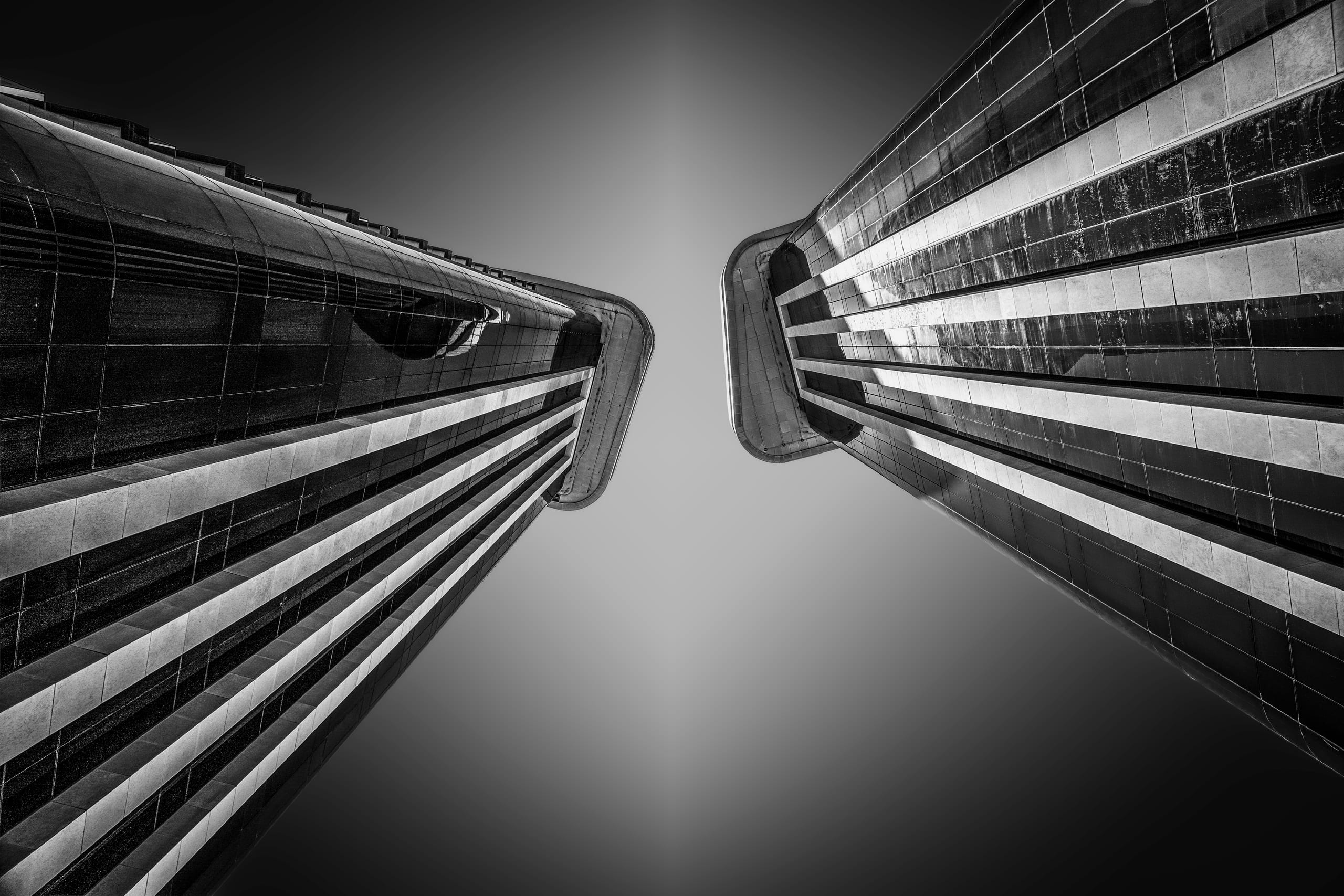 Low-angle monochrome photograph looking straight up between two tall modern buildings with gridded glass facades, creating a V-shaped framing against a dark sky.