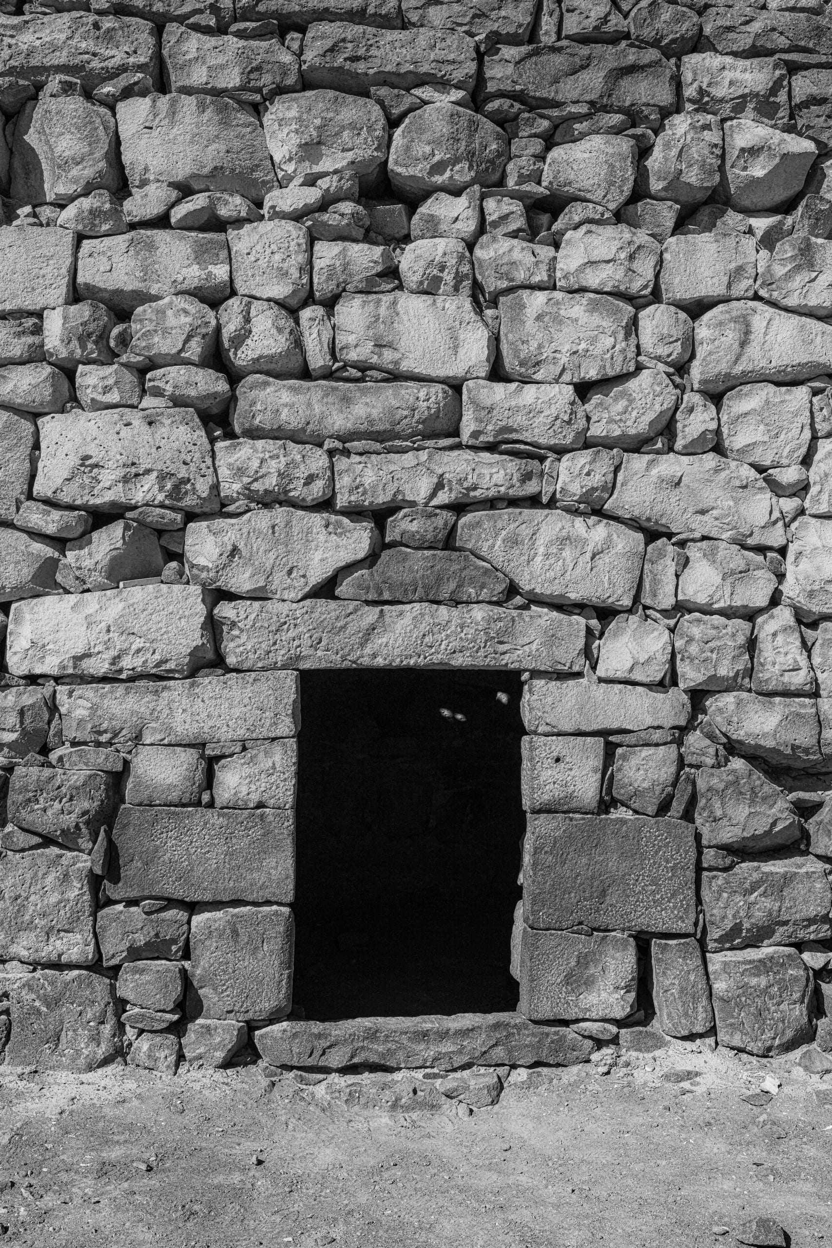 Monochrome architectural photograph of a thick stone wall constructed from irregular ashlar blocks, featuring a dark, centered rectangular doorway at the base.