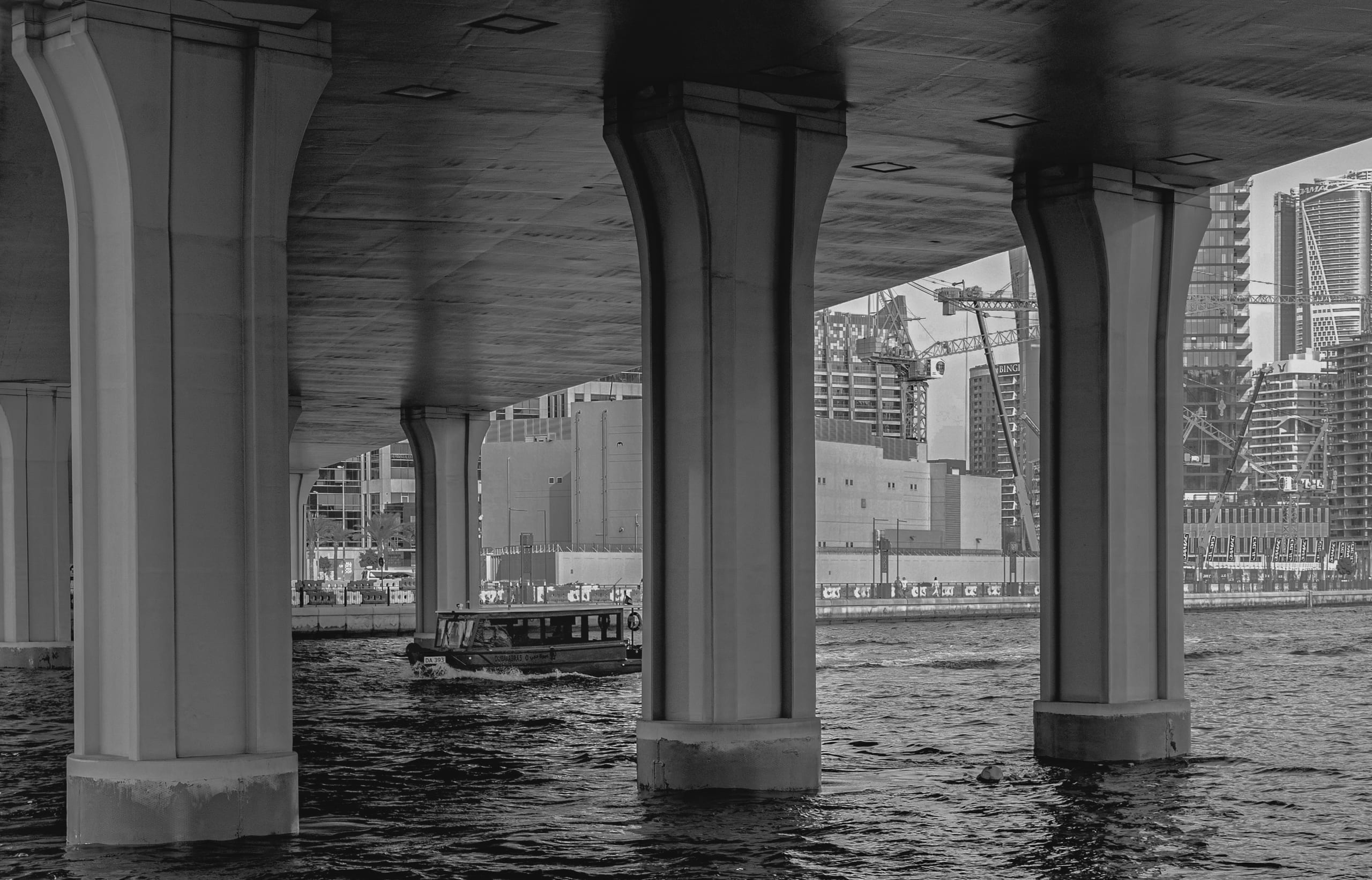 Monochrome architectural photograph showing a boat moving across water under the large vertical piers of a concrete bridge. In the background, city high-rises and construction cranes are visible.