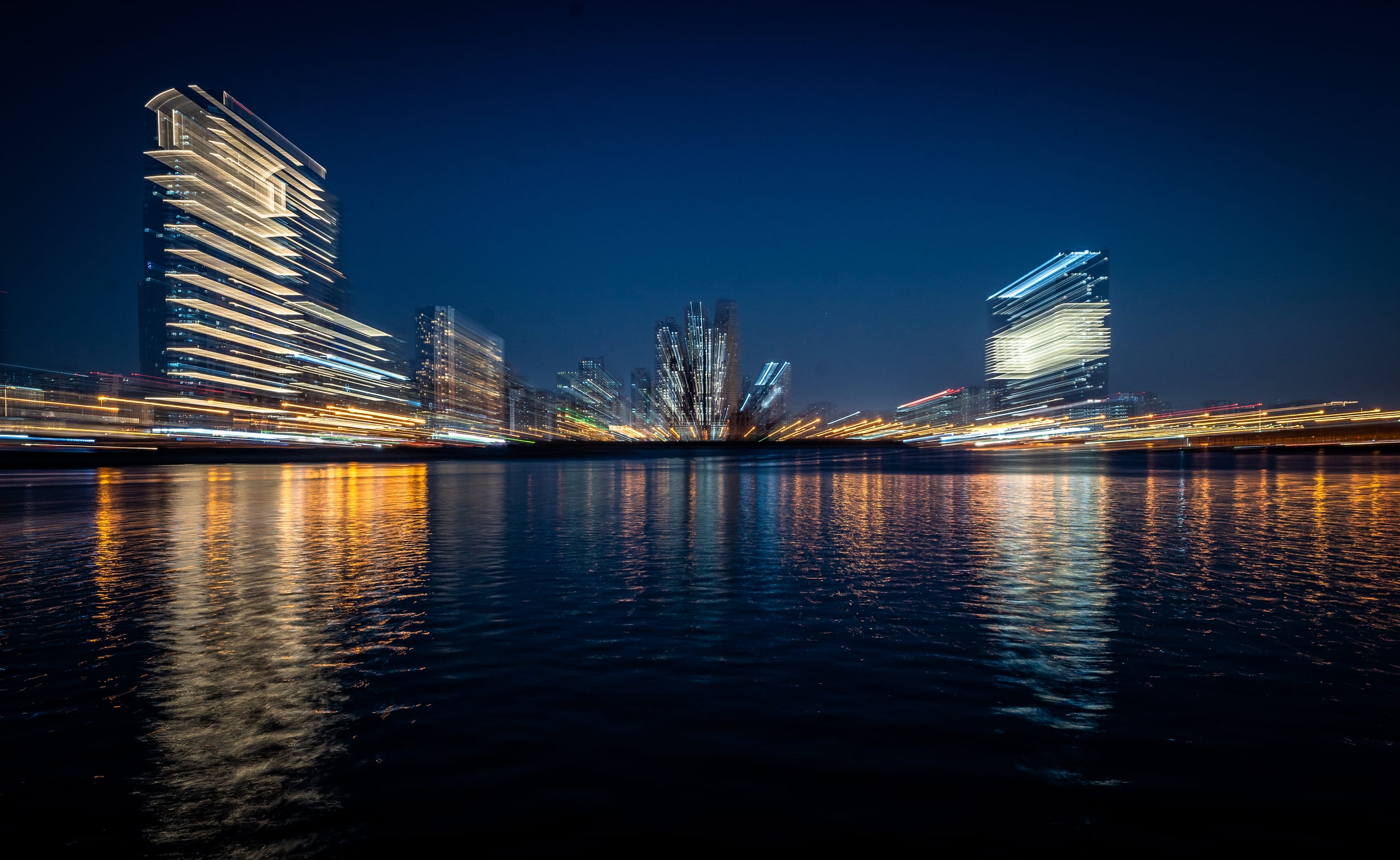 Long-exposure night photograph of illuminated skyscrapers across a body of water, featuring horizontal light streaks and blurred reflections on the water surface.