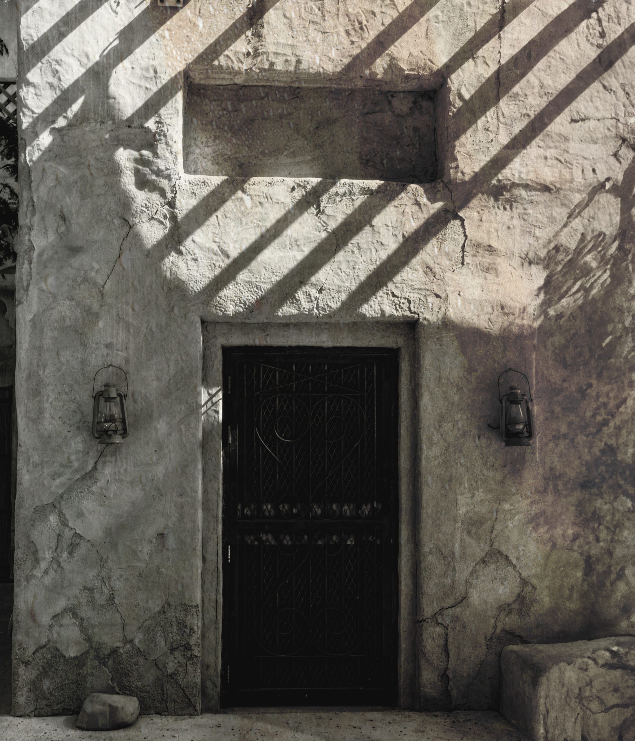 A weathered textured plaster wall with a centered dark door, flanked by two wall-mounted lanterns, crossed by sharp diagonal shadow bars from an overhead pergola.