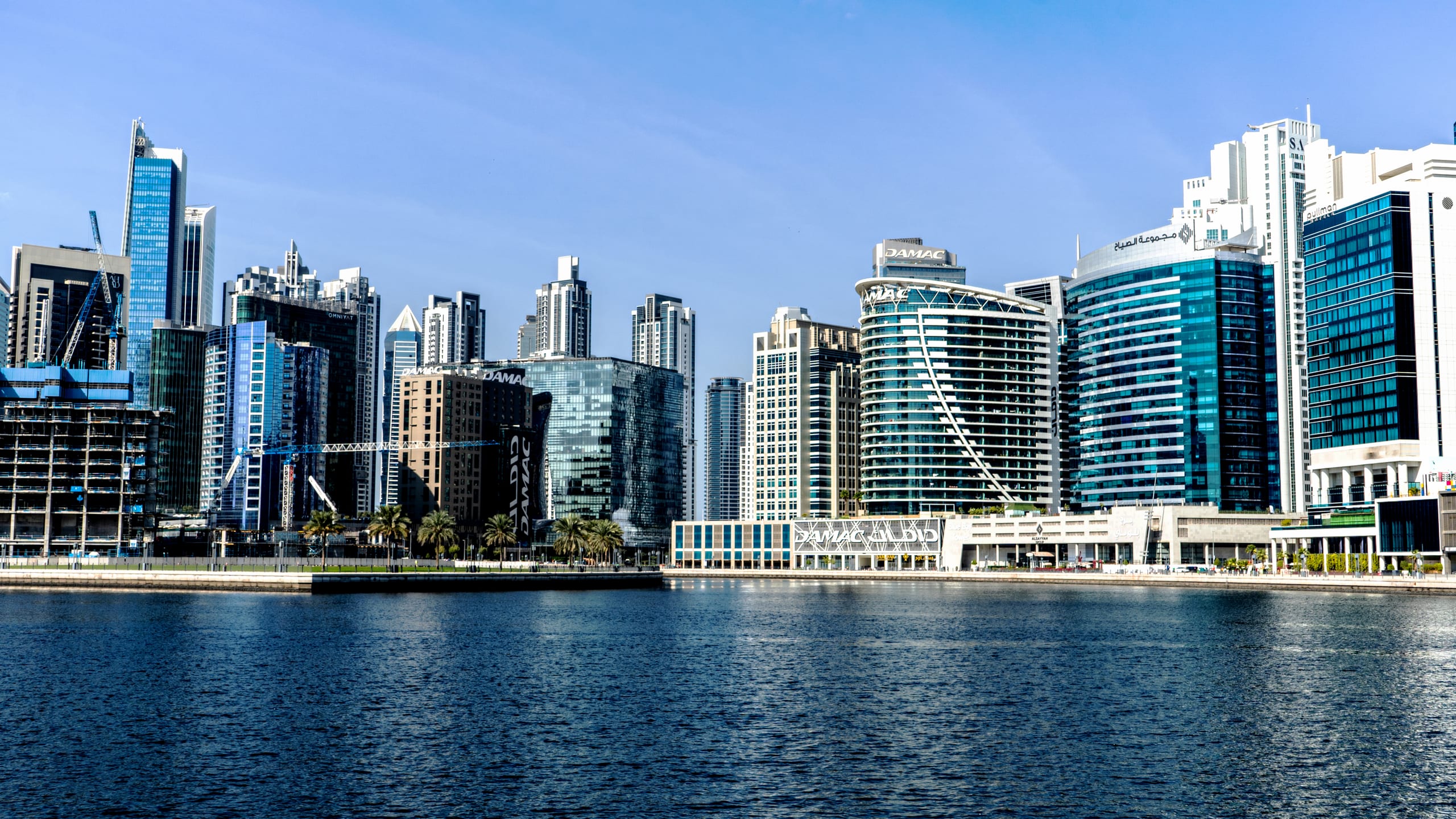 Wide-angle exterior photograph of a modern skyline with numerous skyscrapers along a waterfront, featuring blue glass facades, cranes, and a calm sea in the foreground under a clear sky.