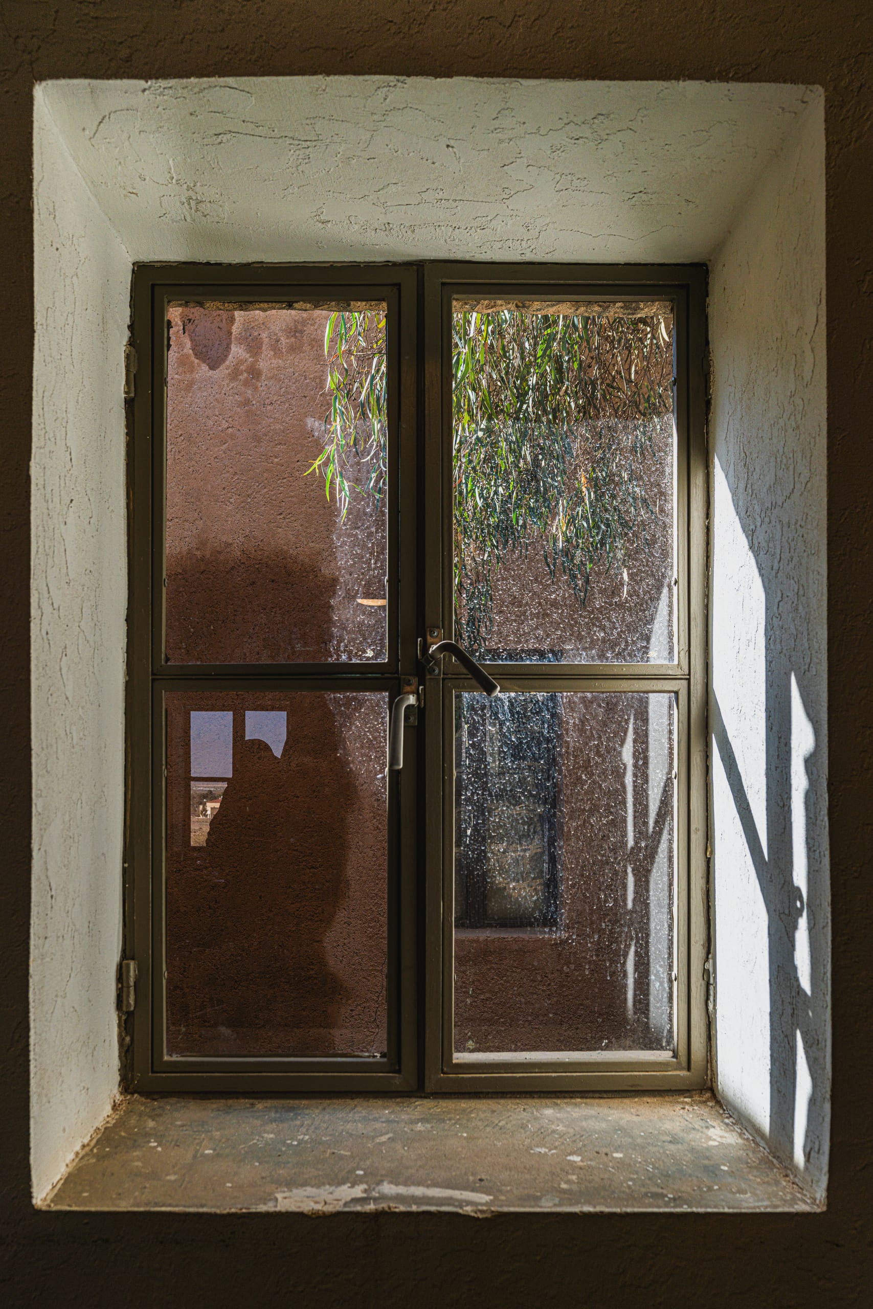 A weathered double-paned window recessed in a thick white plaster wall, showing dusty glass panes reflecting an exterior wall and hanging green foliage in natural light.