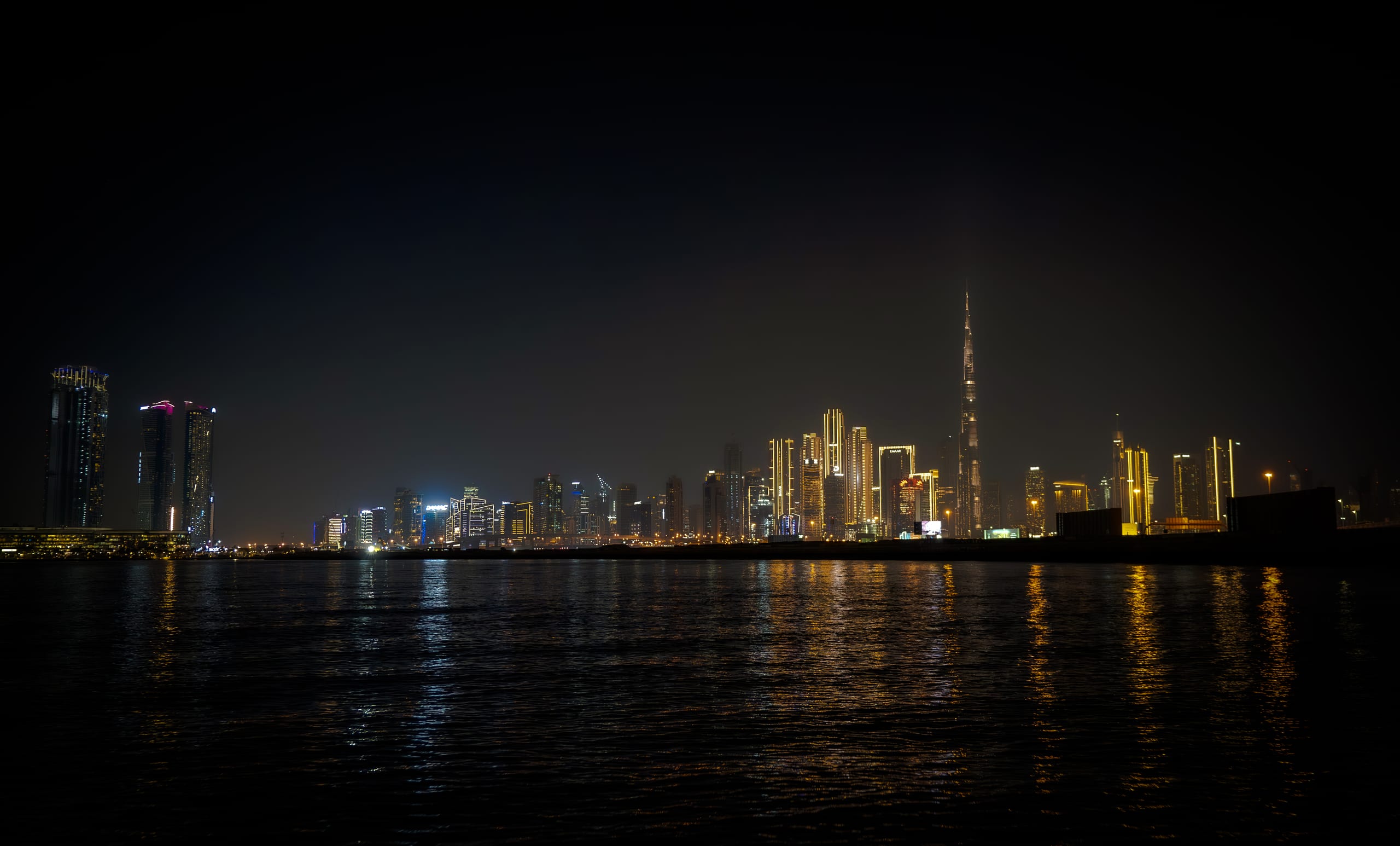 Distant illuminated skyscrapers and a prominent spire at night, viewed across dark water reflecting the city lights.