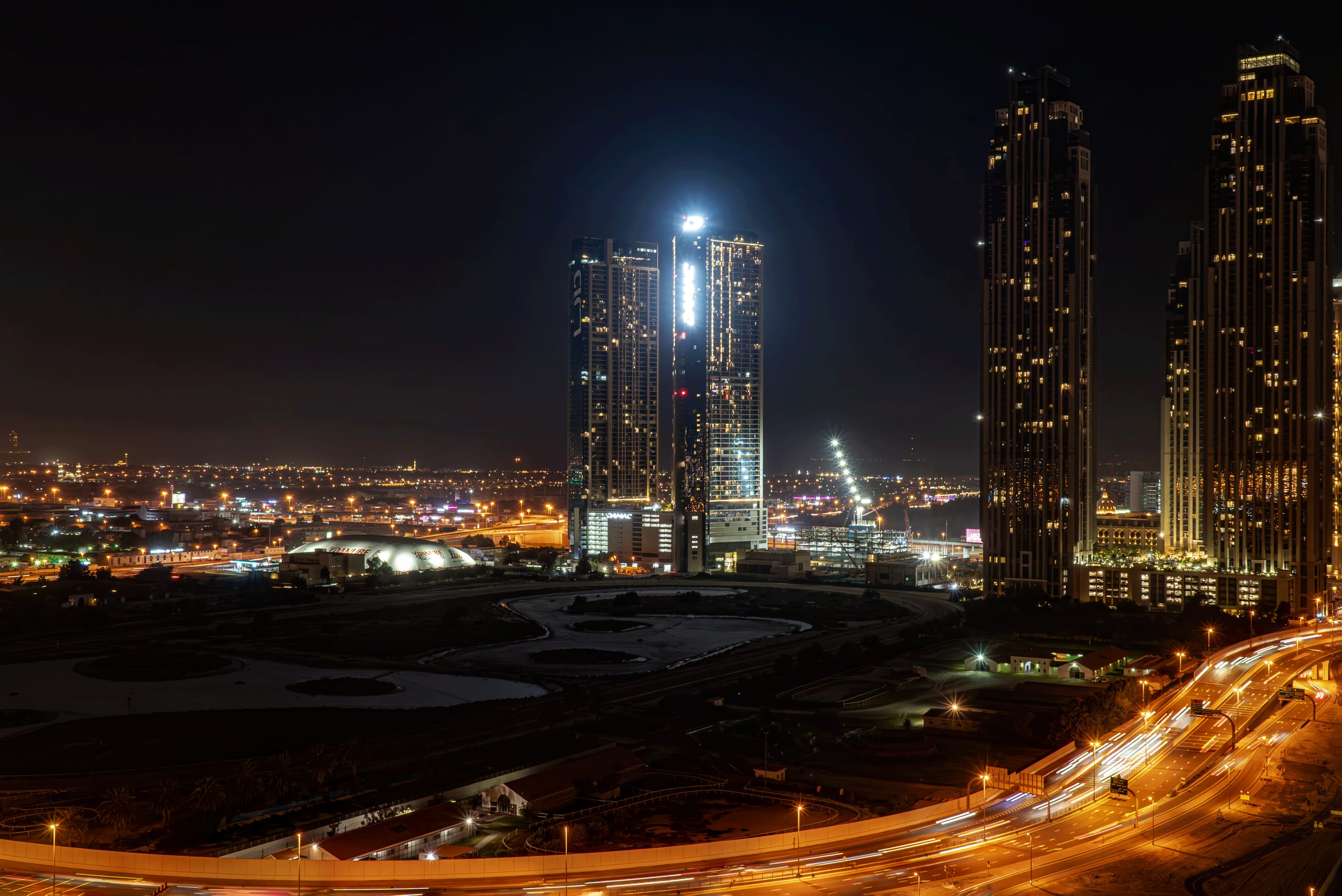 High-angle night view of tall illuminated skyscrapers and a brightly lit curved highway with light trails from vehicle traffic, overlooking an urban landscape in Dubai, UAE.