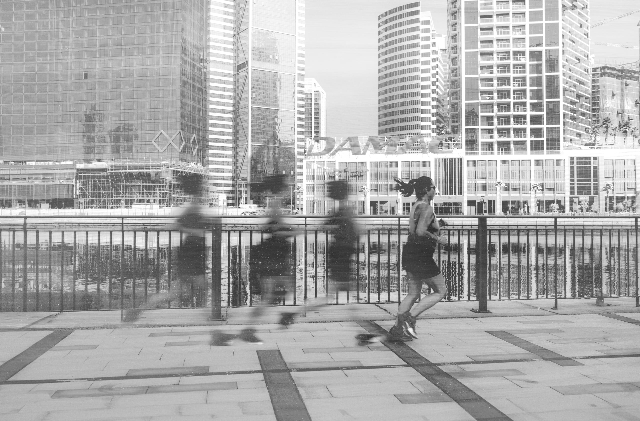 Monochrome image of a female runner in motion along a metal-railed promenade, with blurred multi-exposure ghosts of her figure trailing behind her and dense high-rise skyscrapers in the background.