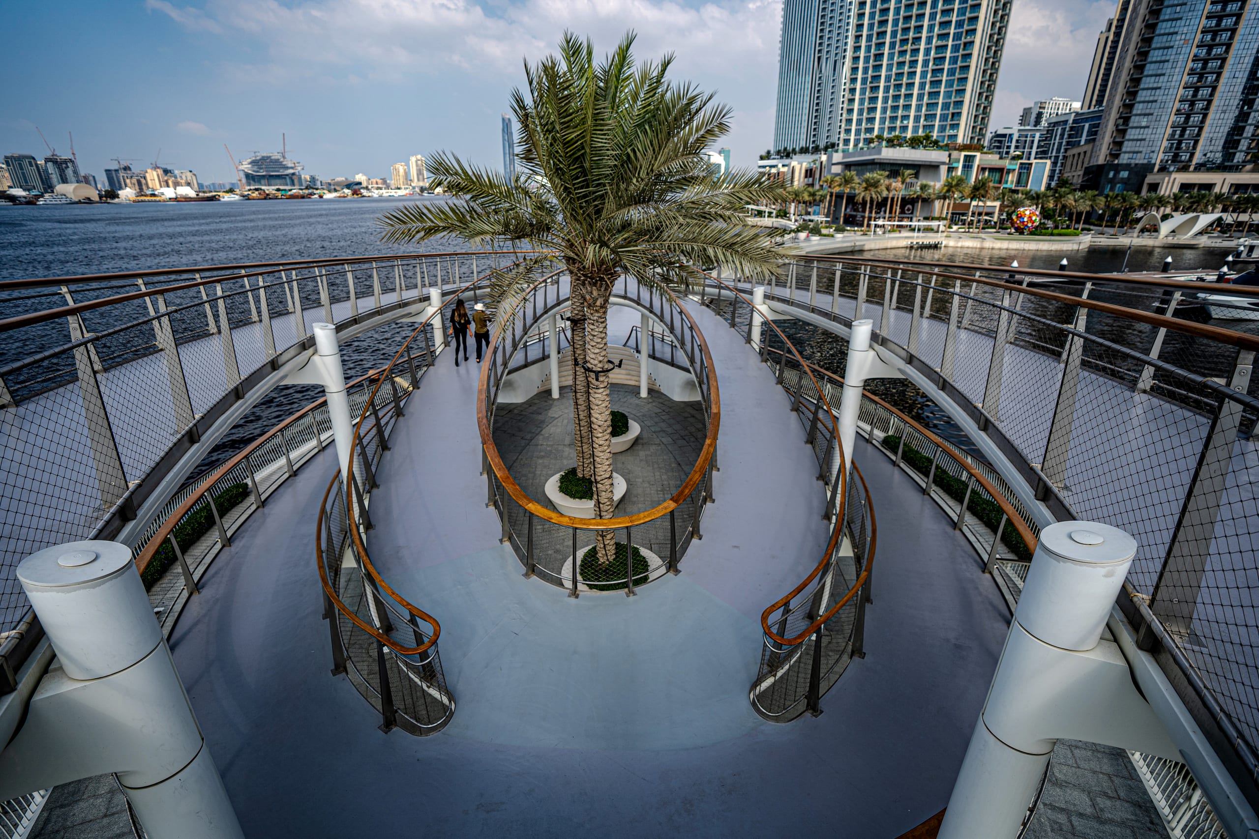 High-angle view of an oval pedestrian bridge with smooth grey pathways, wooden-topped metal railings, and a cluster of palm trees in the center, overlooking a harbor and skyscrapers in Dubai, UAE.