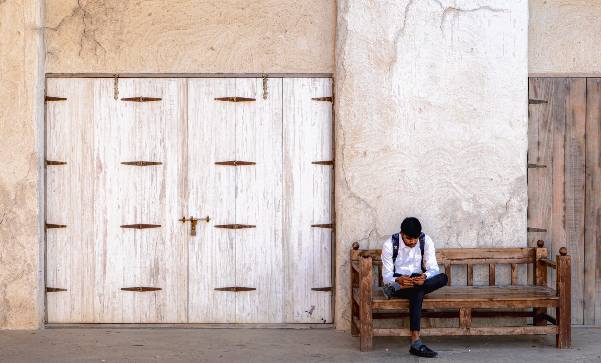 A man in a white shirt and dark trousers sits on a wooden bench against a textured plaster wall, looking down at his mobile phone next to a set of large, white-painted wooden doors.