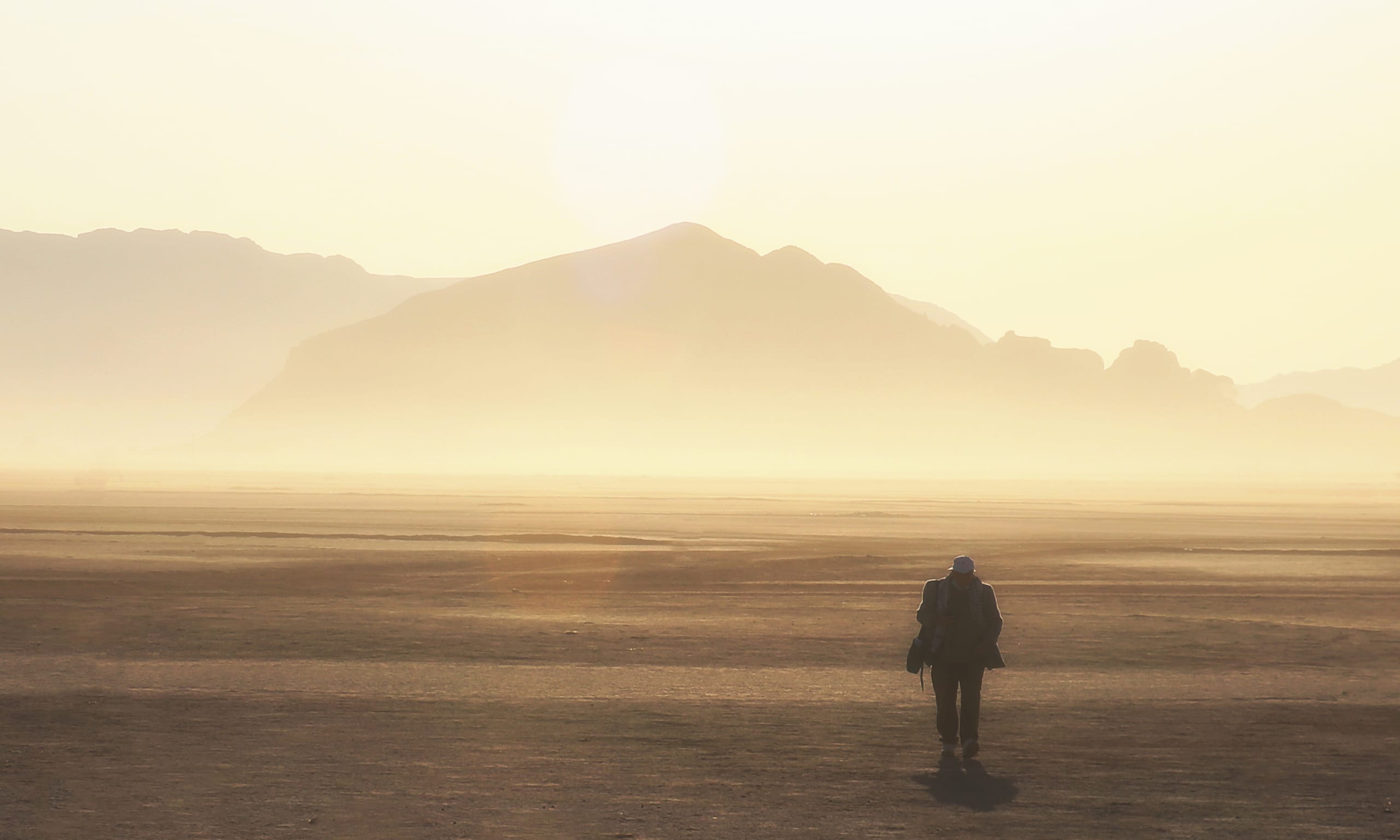 A lone figure walking across a vast, dusty plain towards distant hazy mountains under a bright, diffused sun.