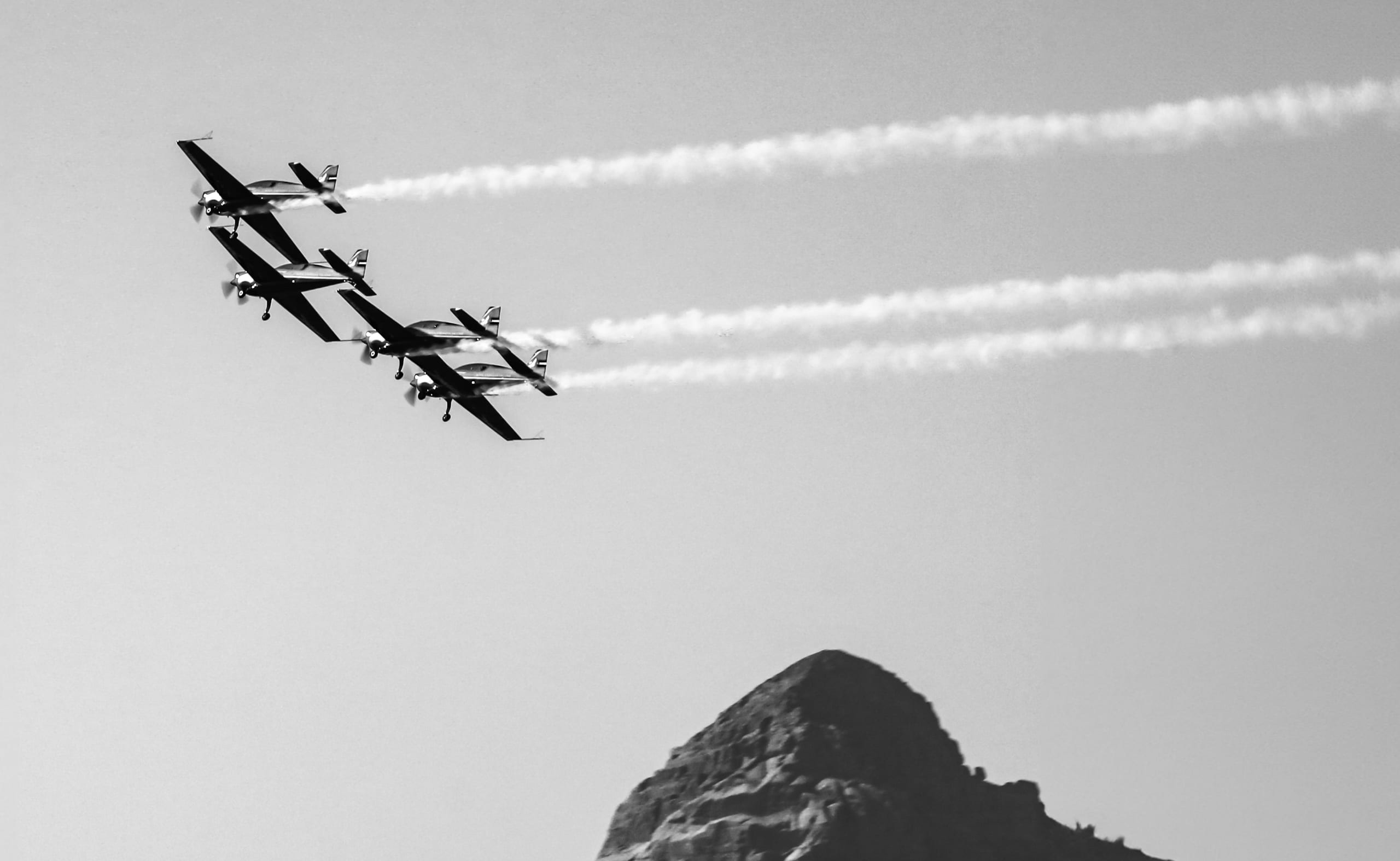 Black-and-white photograph of four propeller planes flying in a diagonal echelon formation, leaving white smoke trails against a pale sky above a rocky mountain peak in the lower right.