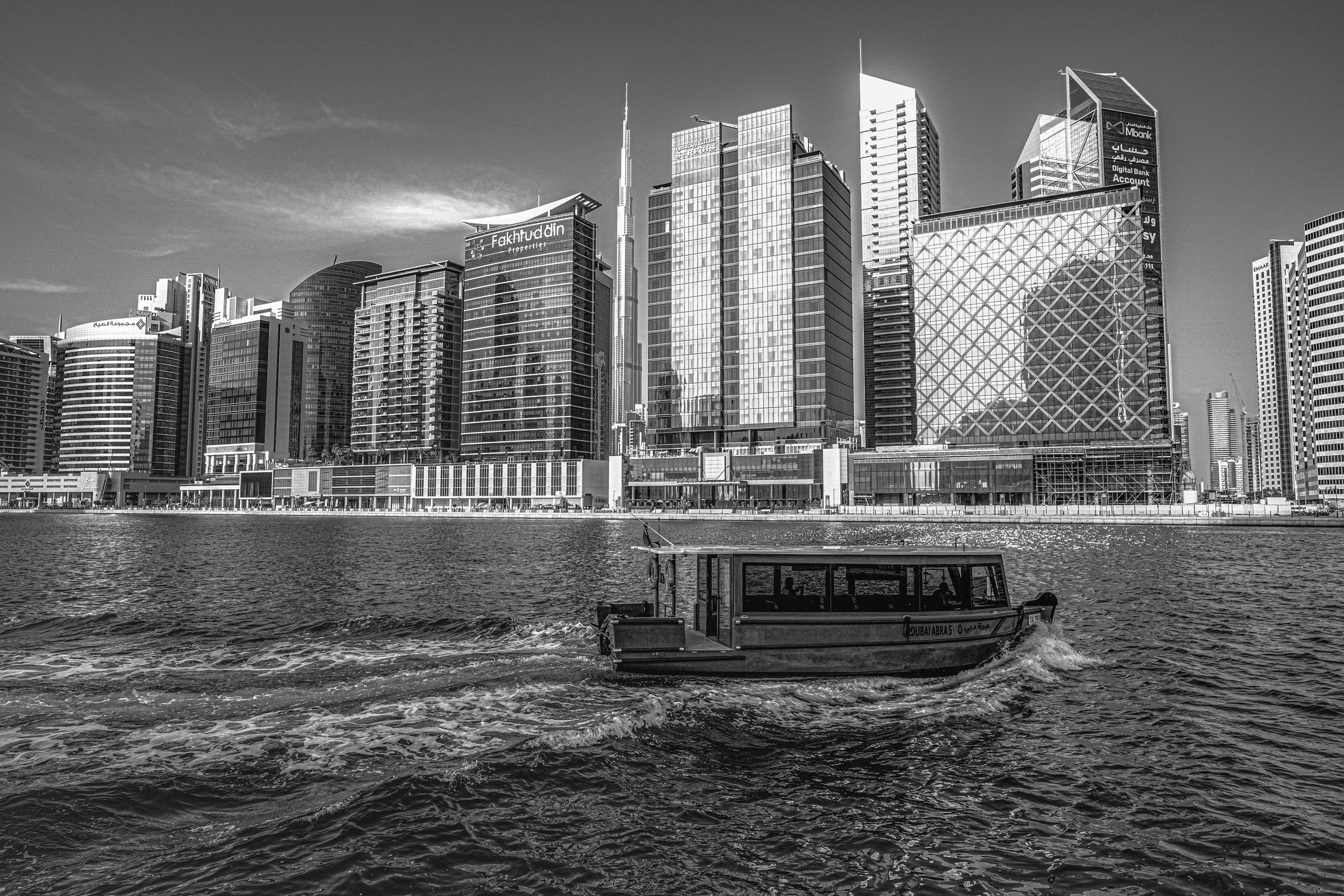A black-and-white photograph of a covered water taxi moving across textured water, with a dense skyline of tall modern high-rise buildings and a slender spire in the background.