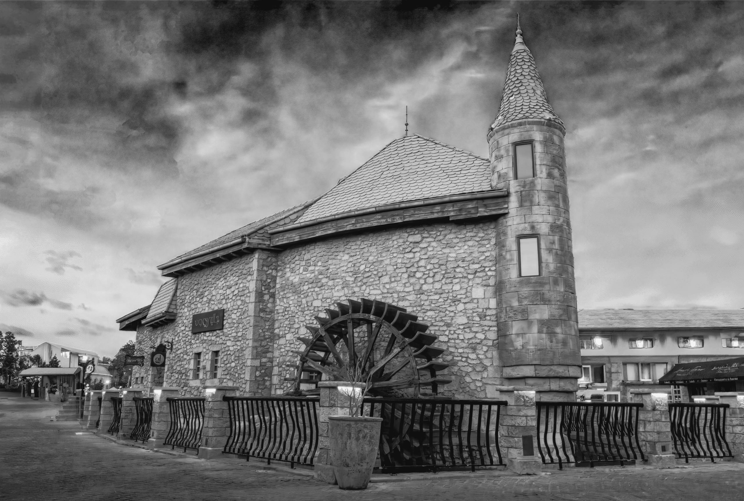Low-angle black-and-white photograph of a stone building with a large wooden water wheel and a circular tower, viewed behind a metal fence under a heavy, cloudy sky.