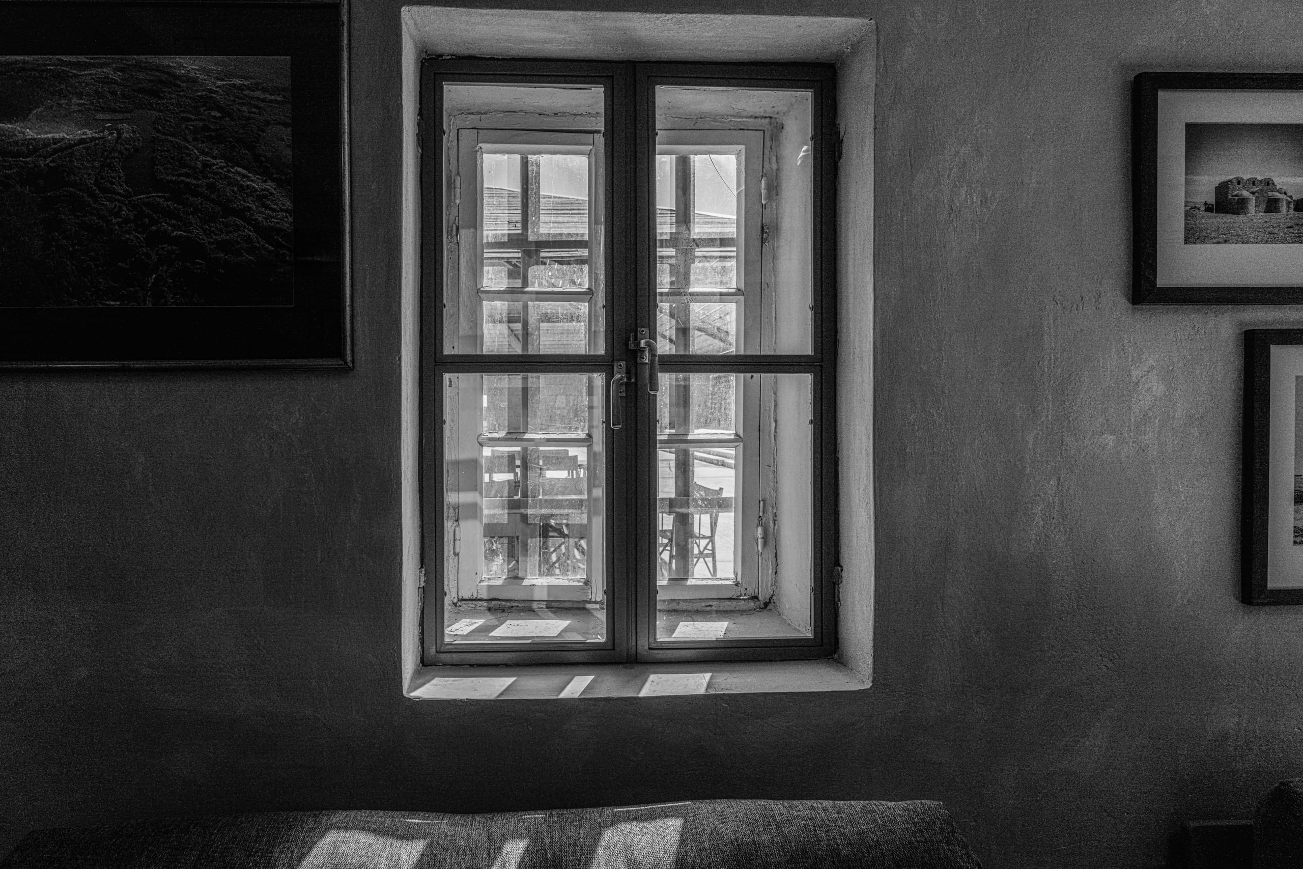 Black and white interior photograph centered on a multi-pane window in a textured wall, flanked by framed artwork, with bright light casting geometric patterns onto a sofa in the foreground.