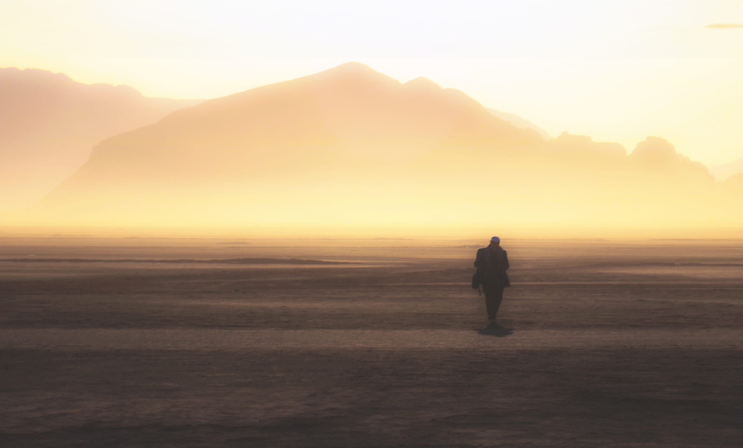 A single person walking away across a vast, flat landscape under a hazy, golden sky with distant mountains silhouetted against the light.