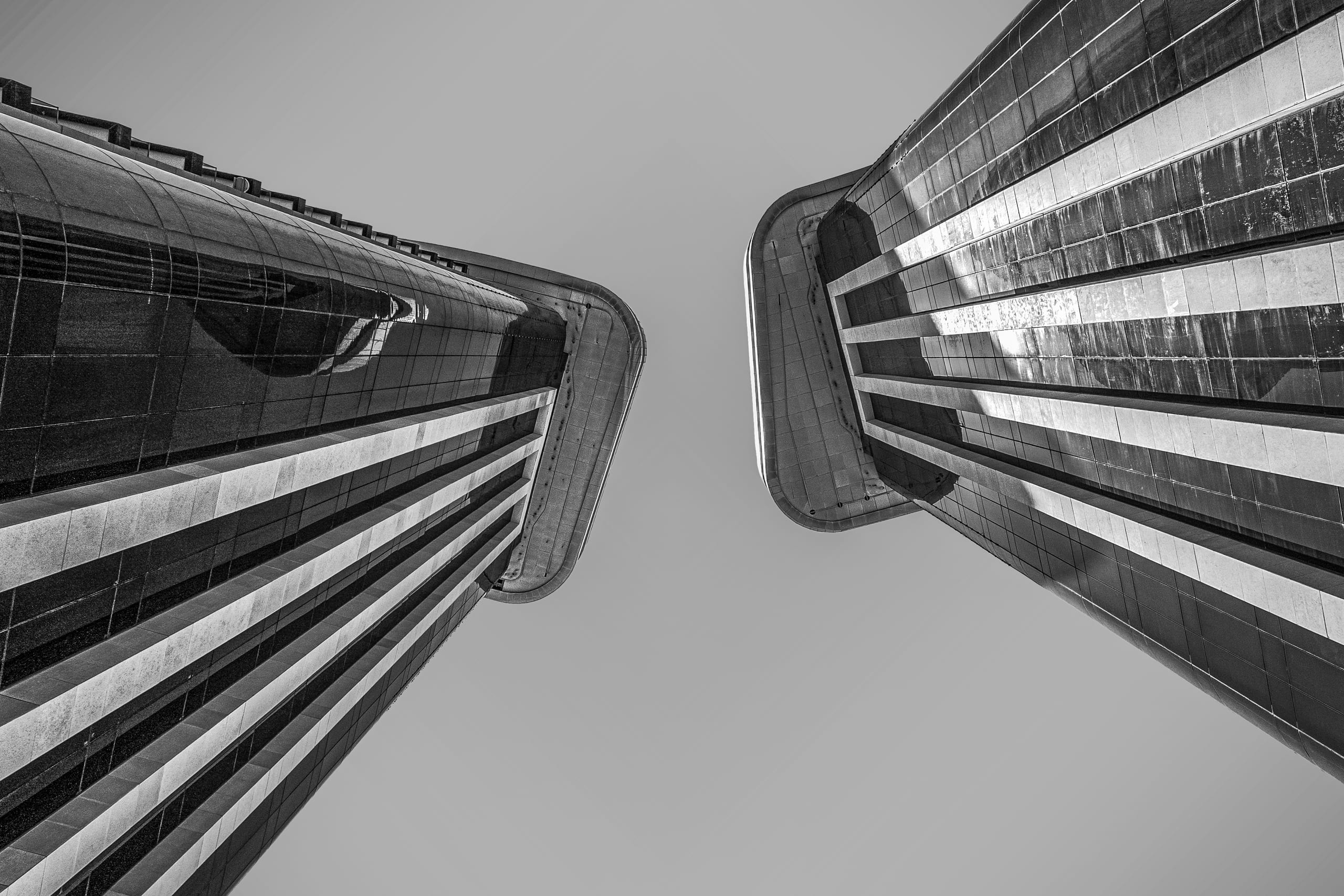 A low-angle monochrome photograph looking straight up at two tall, modern skyscraper facades that curve inward toward each other against an overcast sky.