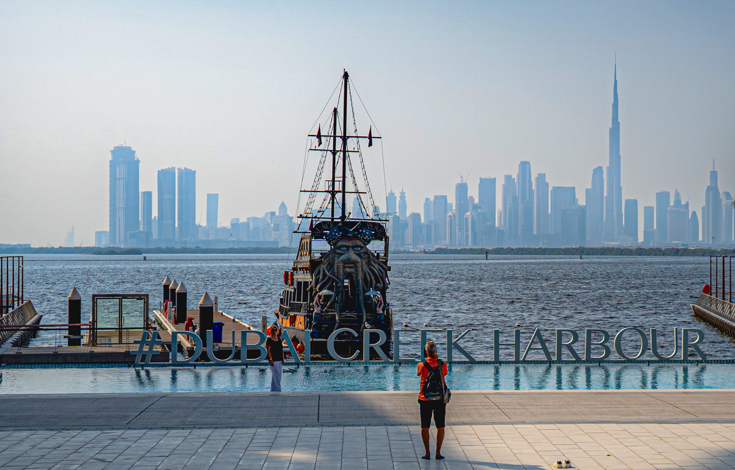 A person viewed from behind stands on a tiled waterfront plaza, facing a large, ornate ship with a carved figurehead and tall masts, with a hazy modern city skyline featuring a very tall skyscraper visible in the background across calm water.