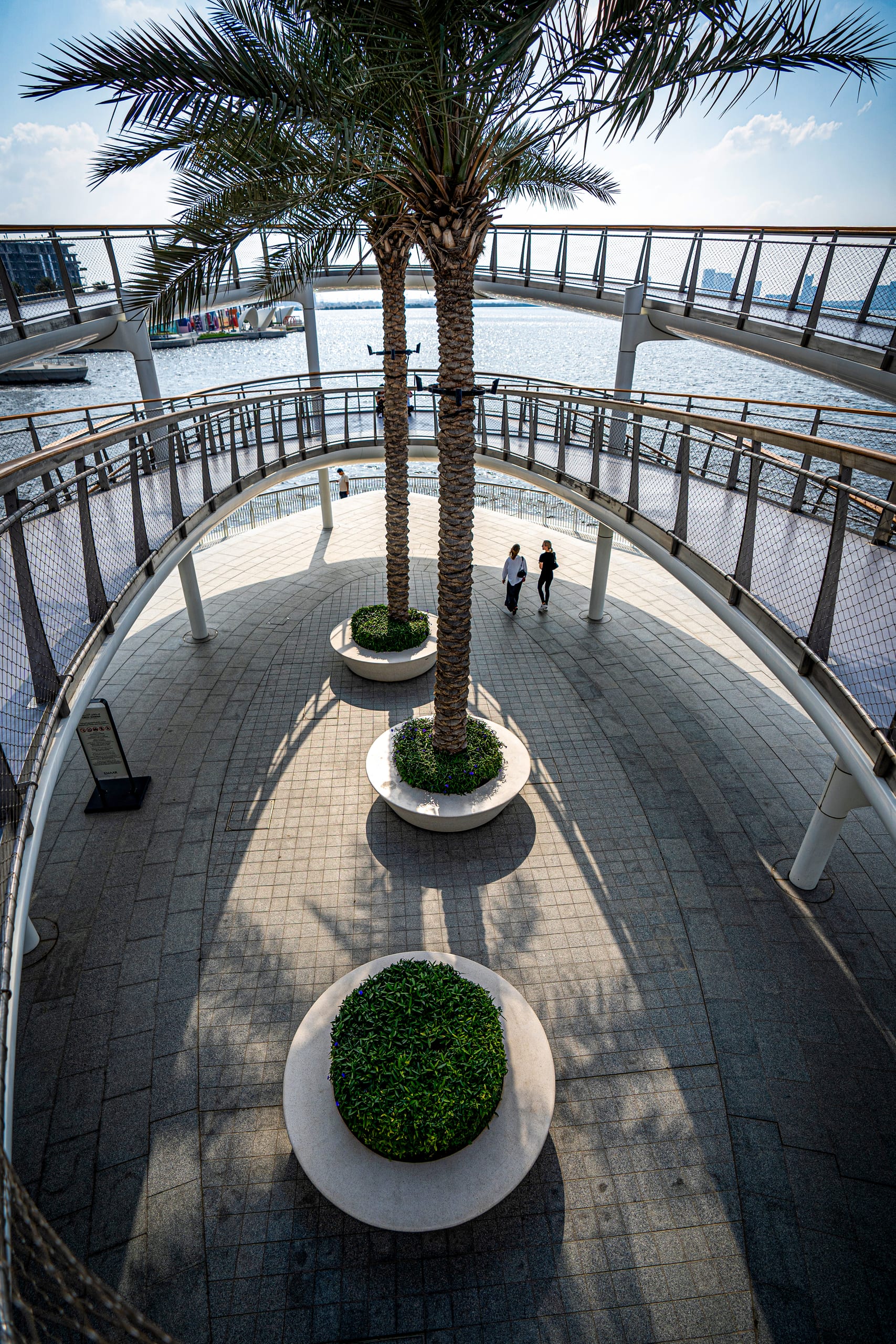 High-angle view of a multi-level pedestrian bridge curving over a paved square with three circular planters and palm trees, with two figures walking below.
