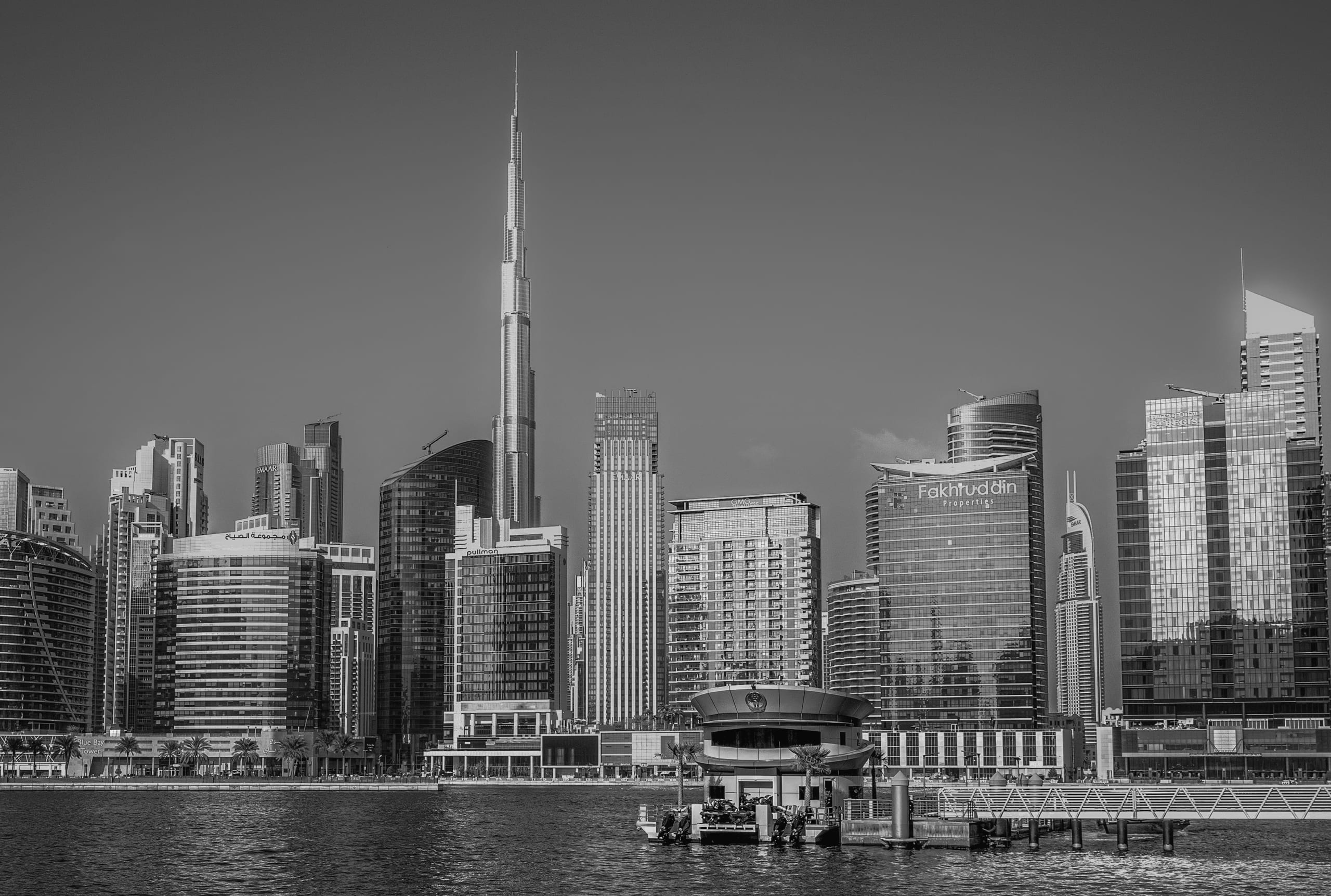 Black and white photograph of a prominent skyscraper rising above surrounding modern high-rises and waterfront structures, with water visible in the foreground.