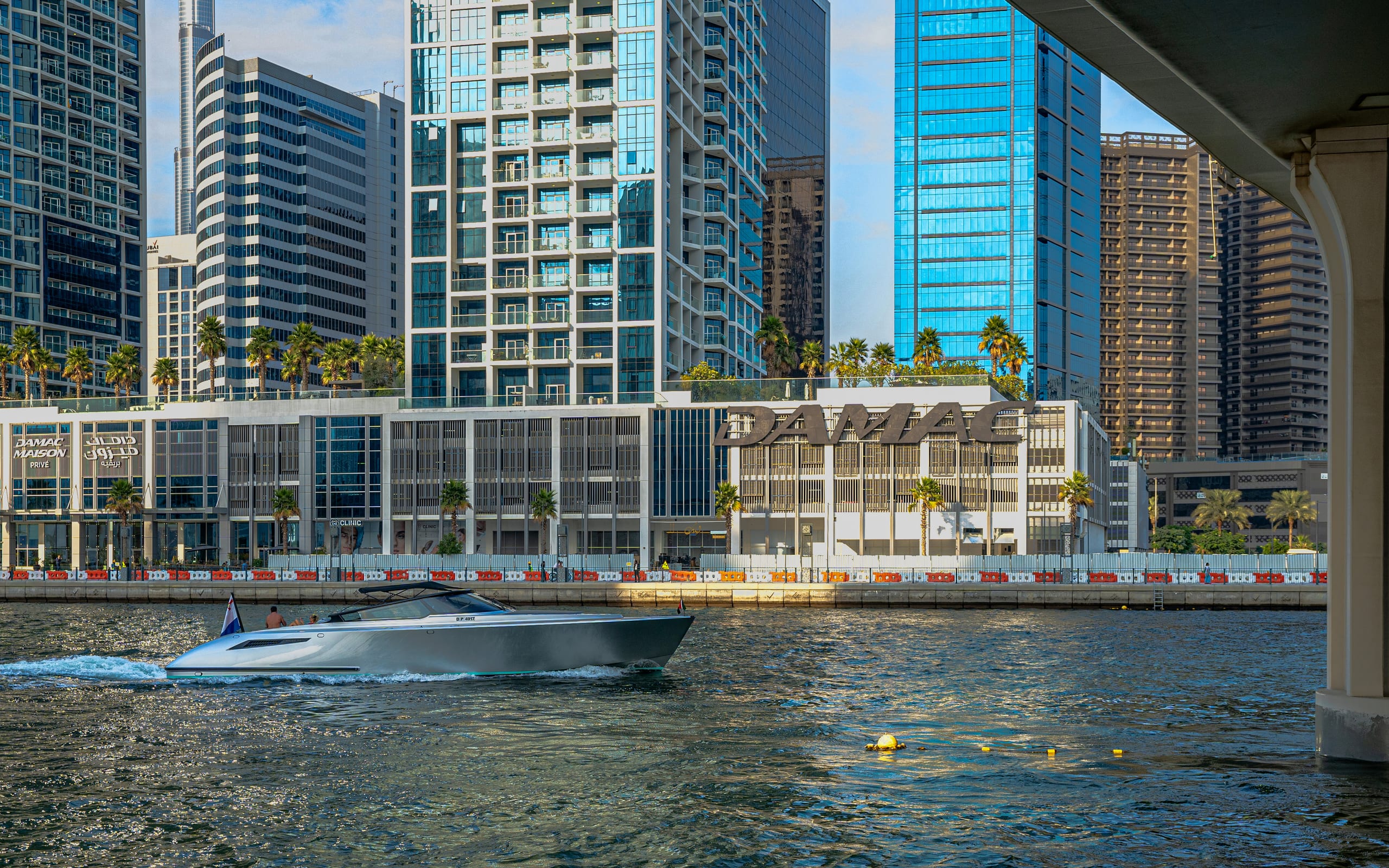 A modern silver motorboat moving left to right across a waterway, framed by towering skyscrapers and a concrete bridge pillar in the foreground right.