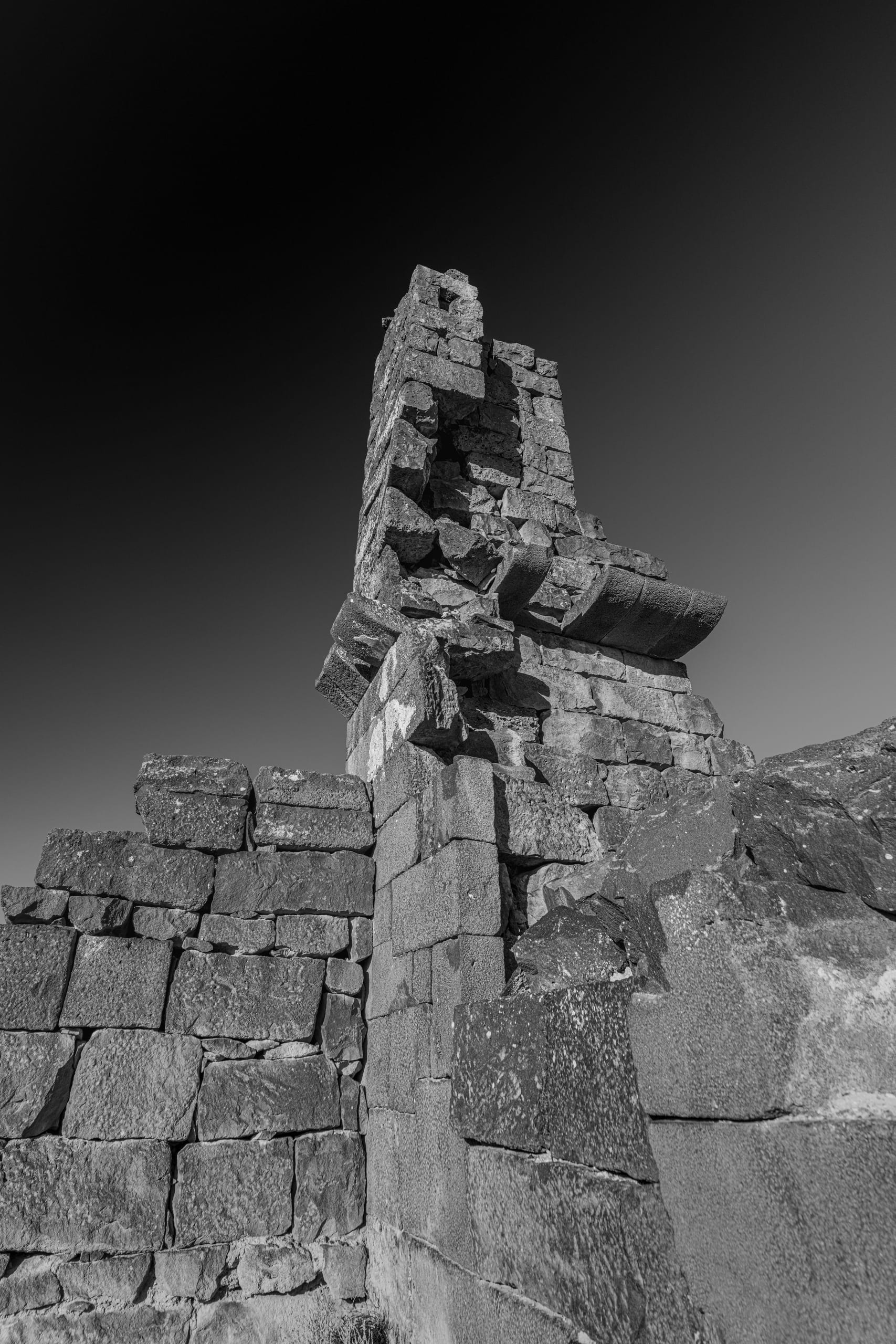 Low-angle black-and-white photograph of ancient stacked stone ruins and a fractured vertical column standing against a gradient sky.