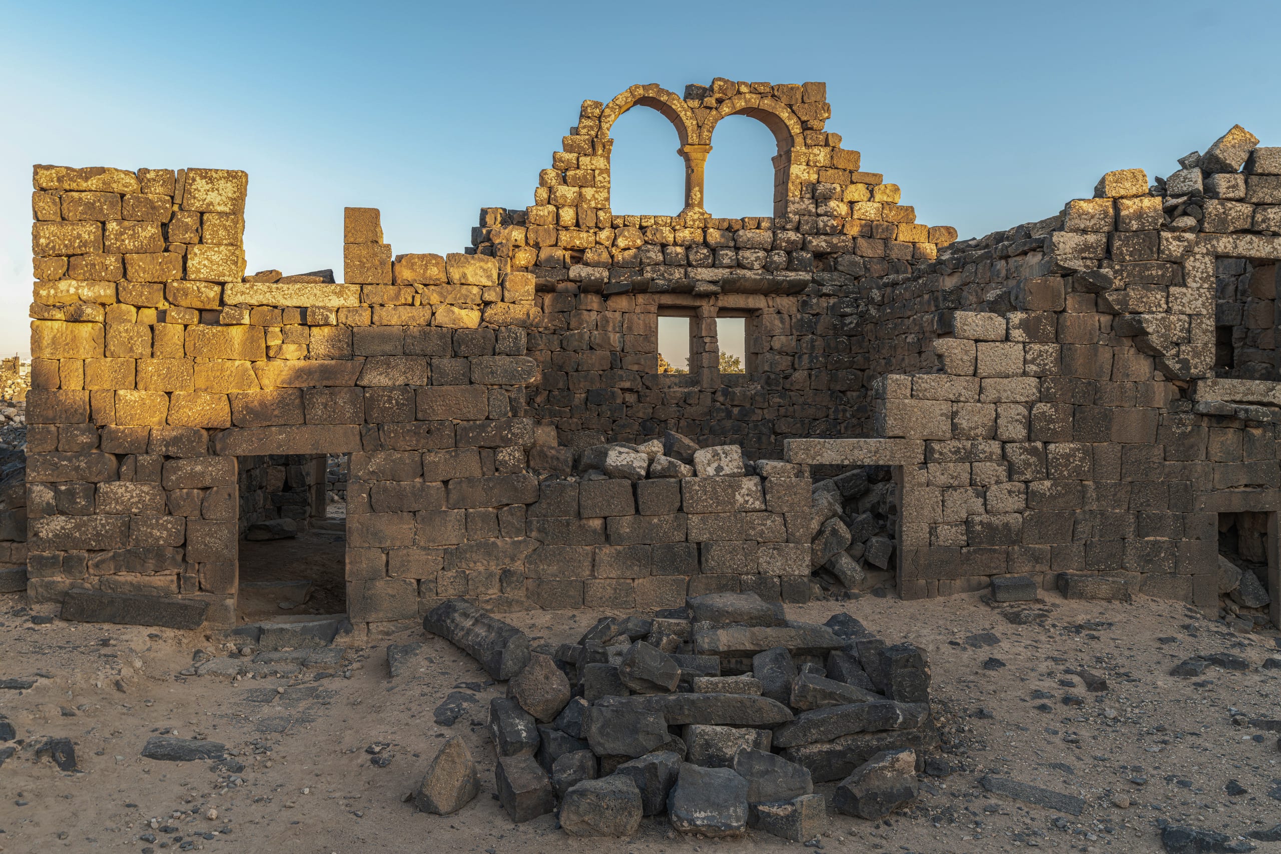 Front view of ancient stone ruins constructed from dark rectangular blocks under a clear blue sky, featuring double upper arches, rectangular lower windows, doorways, and fallen rubble on the ground.