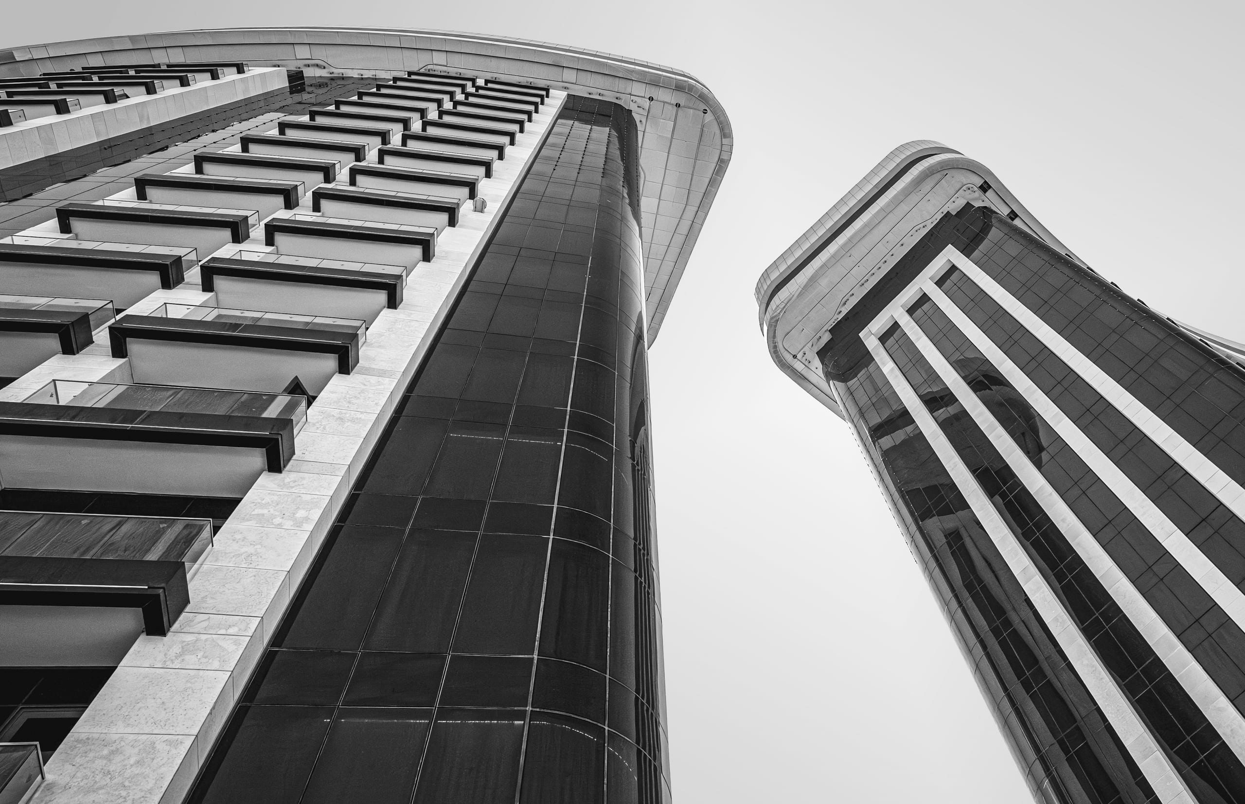A low-angle black-and-white photograph of two tall modern high-rise buildings, showing dark glass panels, rows of balconies, and curved architectural lines against a plain sky.