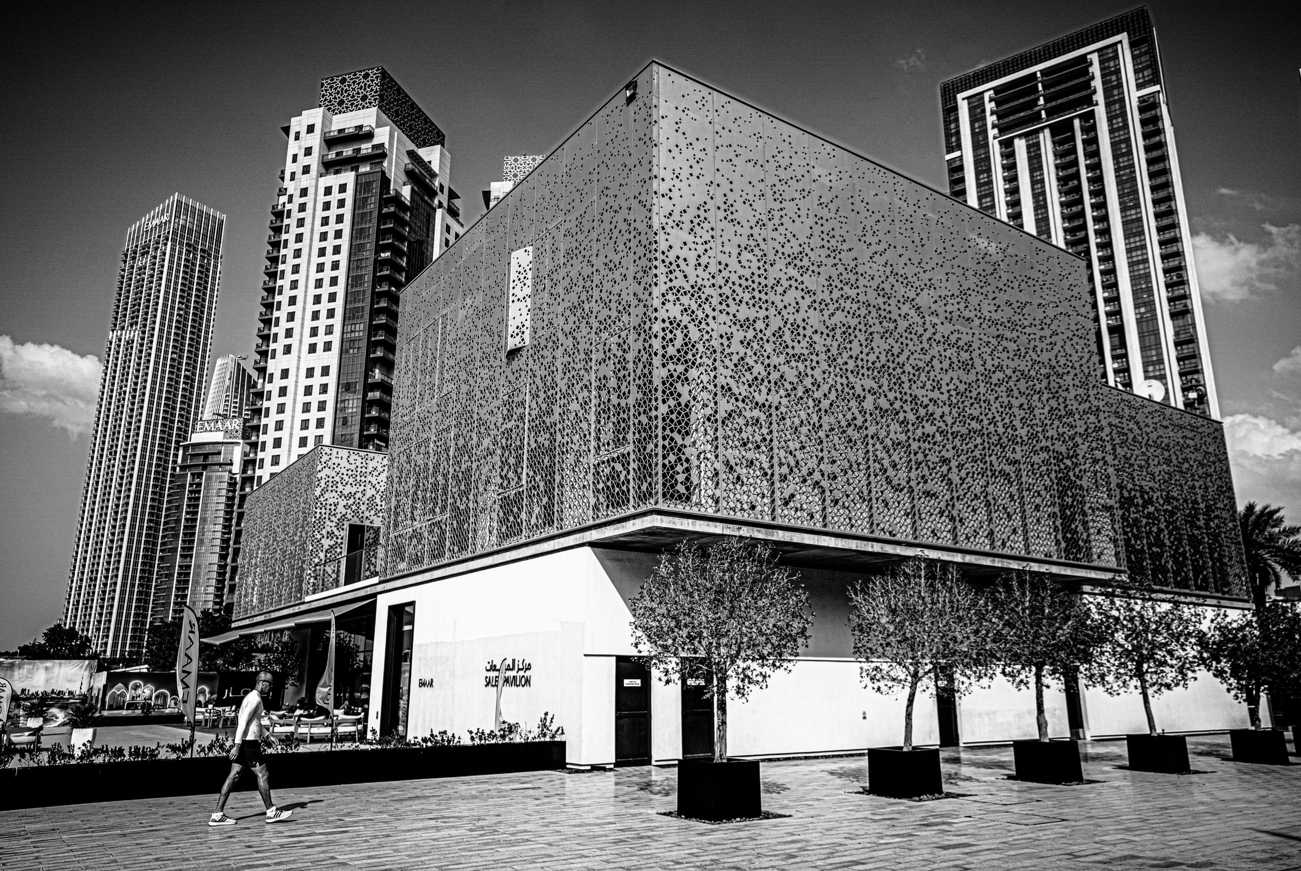 Monochrome architectural photograph showing a lone person walking past a modern pavilion with a perforated facade, set against towering skyscrapers in Dubai, UAE.