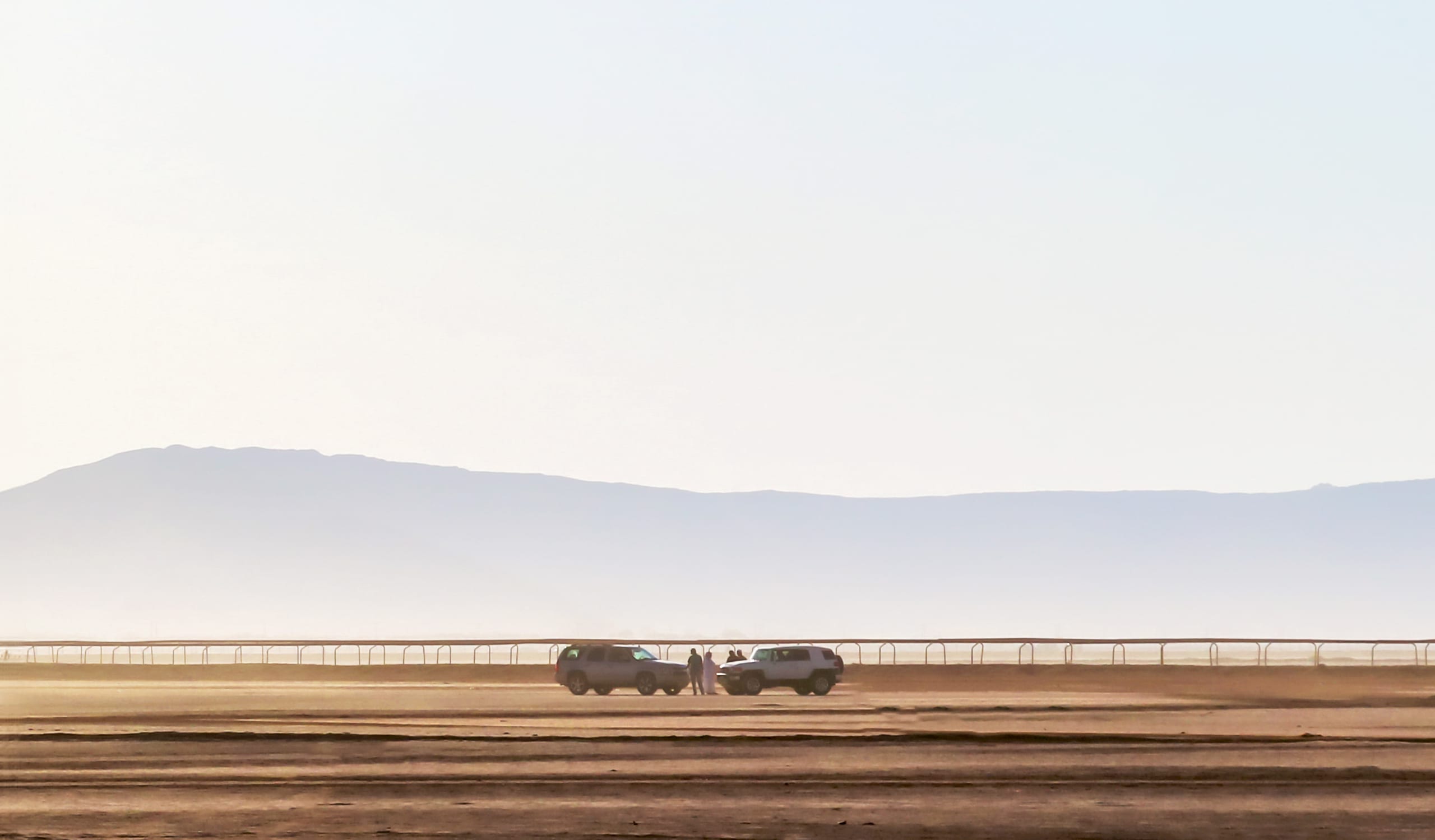 Wide-angle view of a vast desert expanse with two parked SUVs and several small human figures standing between them near a long horizontal fence, backed by faint hazy mountains and a pale sky.