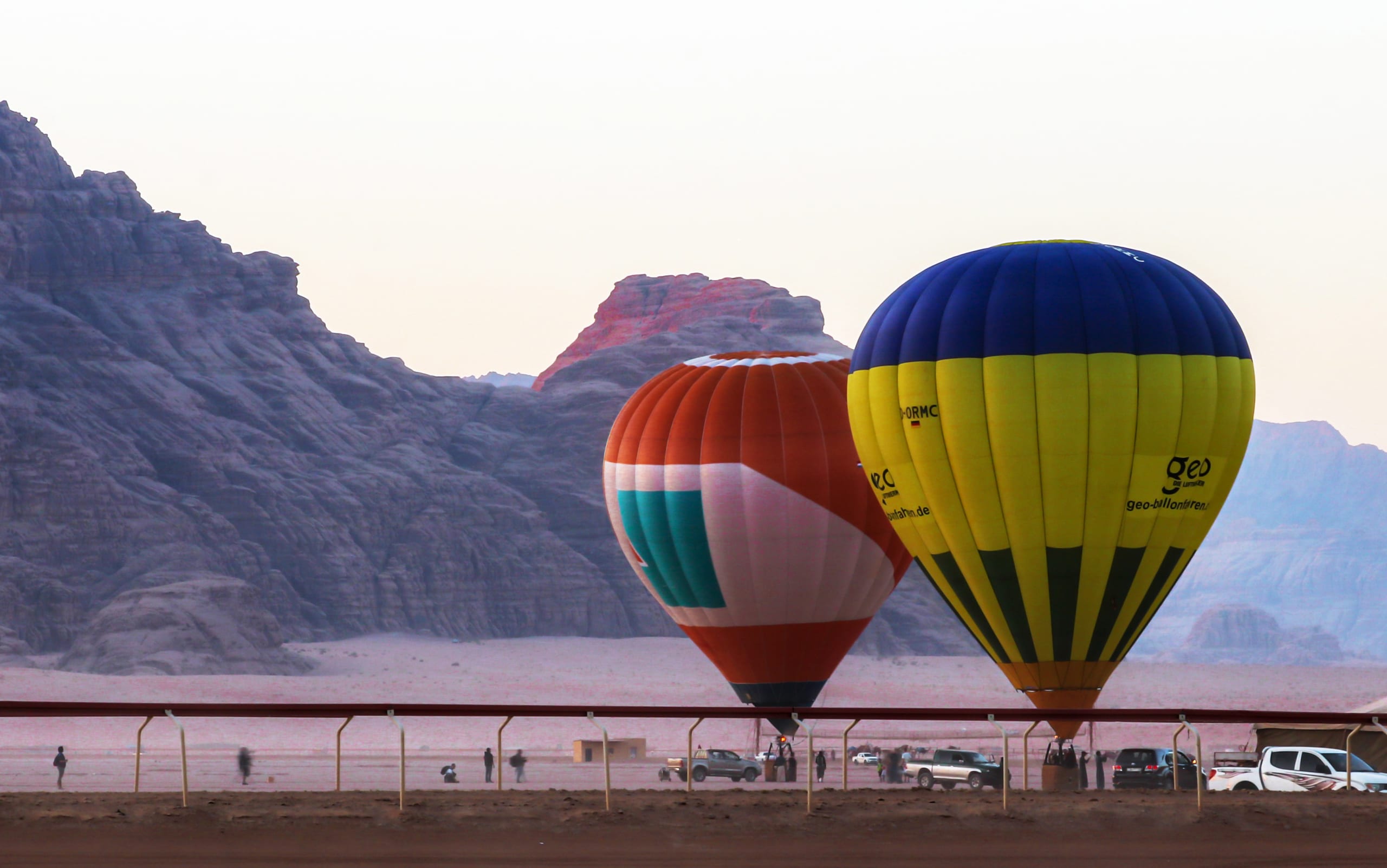 Two hot air balloons, one orange-and-white and one yellow-and-blue, stand grounded in a desert landscape with rocky mountains in the background and a horizontal railing in the foreground.