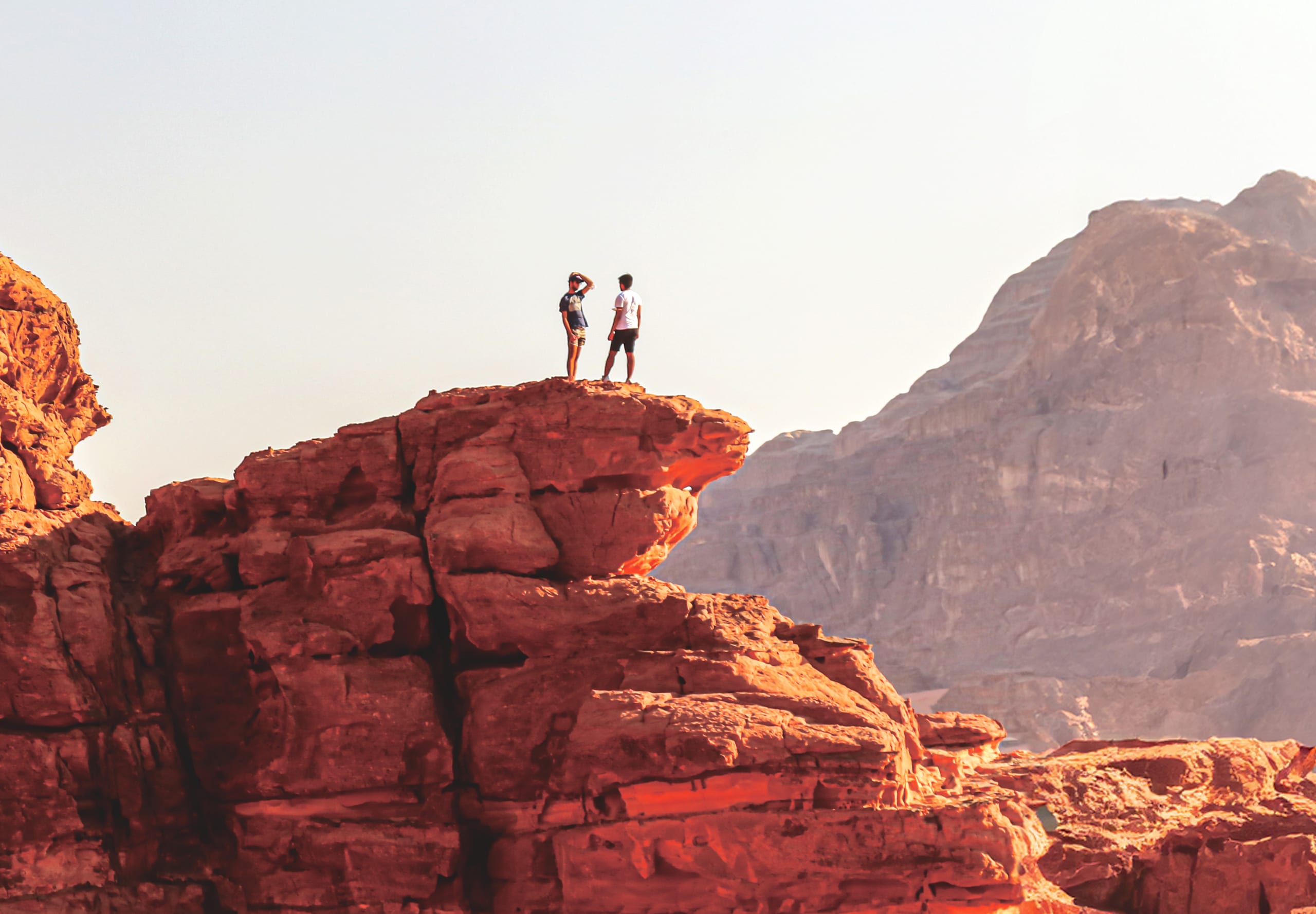 Two people standing on a high red rock plateau, silhouetted against a bright sky, with large rugged mountain slopes in the background.