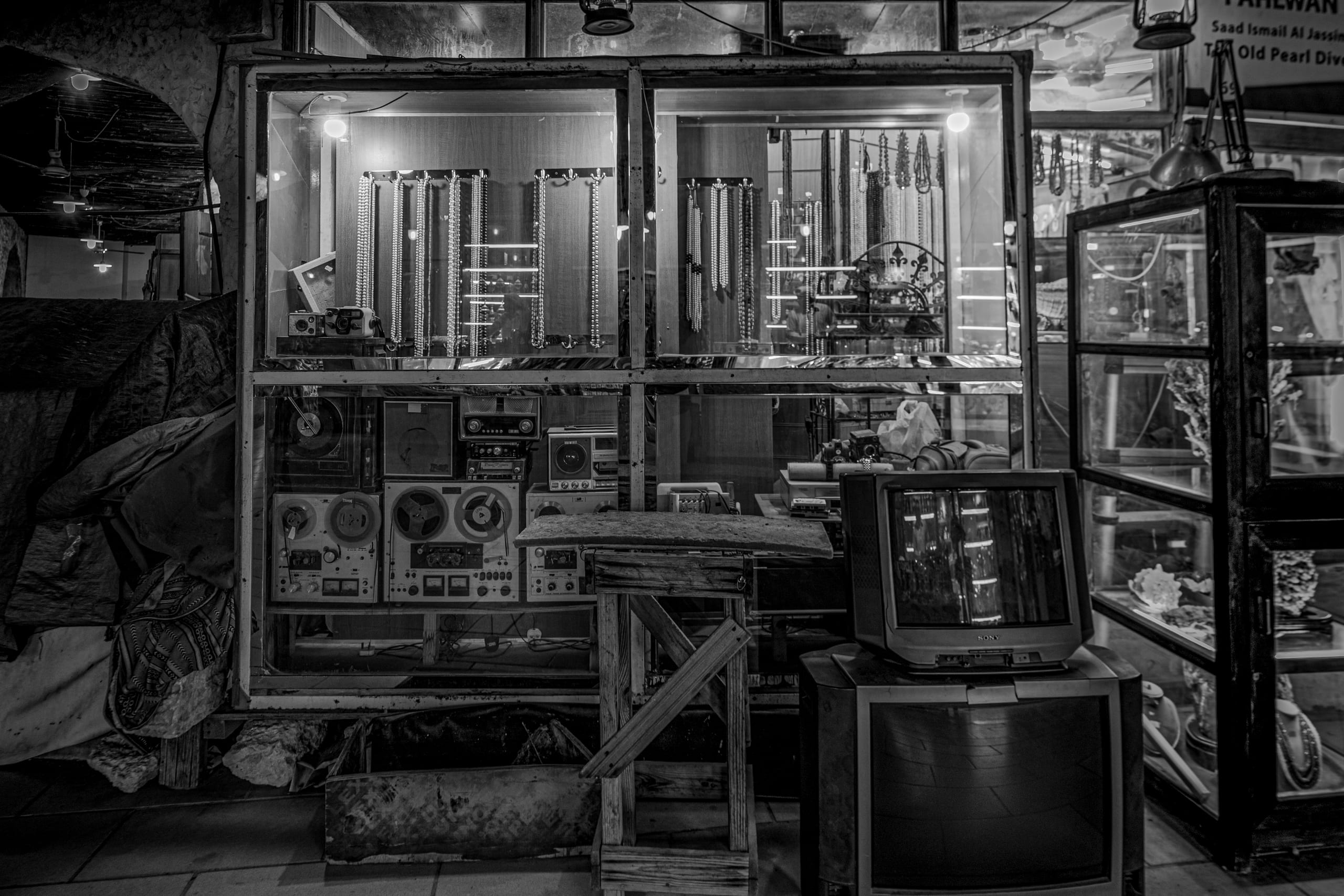 Black-and-white photograph of a shop window densely packed with vintage audio equipment, tape decks, older televisions, and rows of hanging beads and necklaces, with a wooden stool in the foreground.