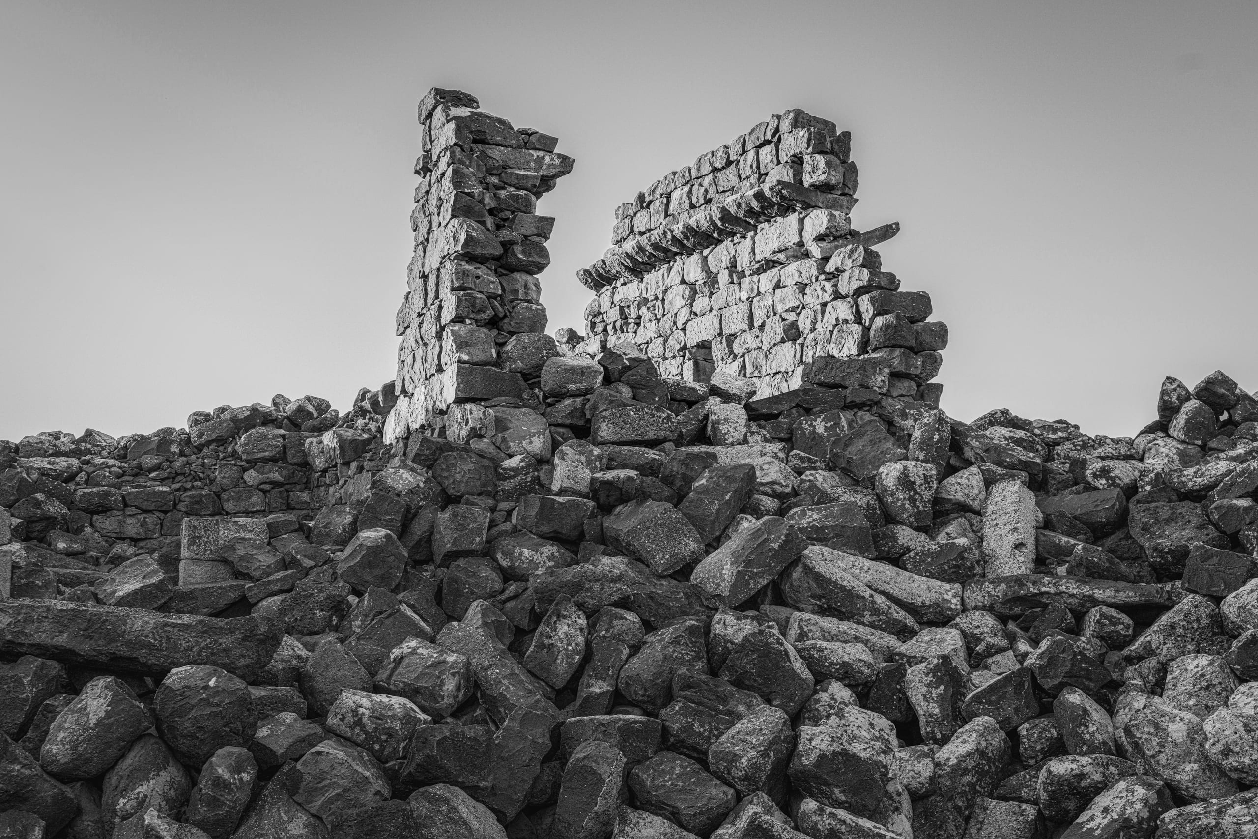 Black-and-white photograph of crumbling ancient stone walls rising behind a vast foreground pile of dark, irregular boulders under a clear sky.