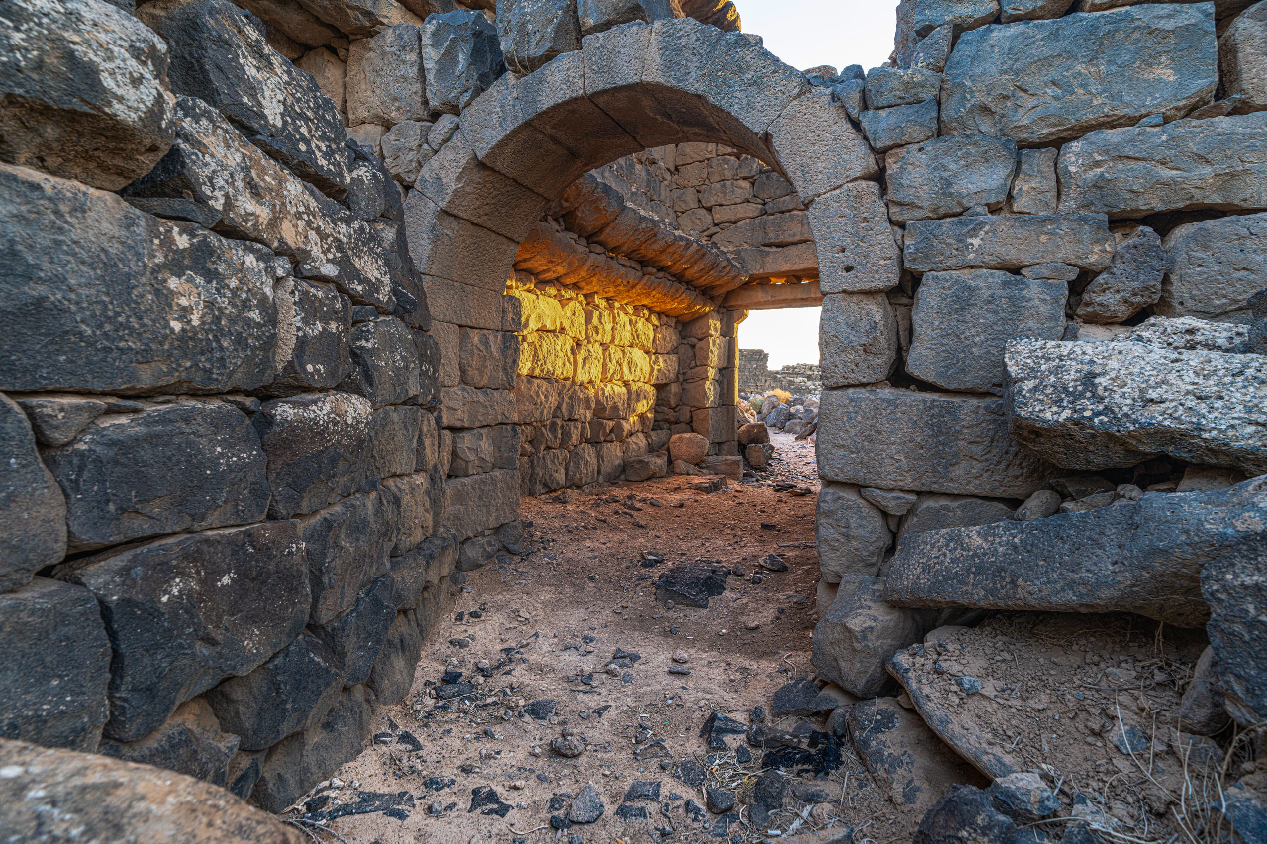 Interior view through a stacked stone archway in an ancient ruin, with bright golden sunset light striking the inner stone walls.