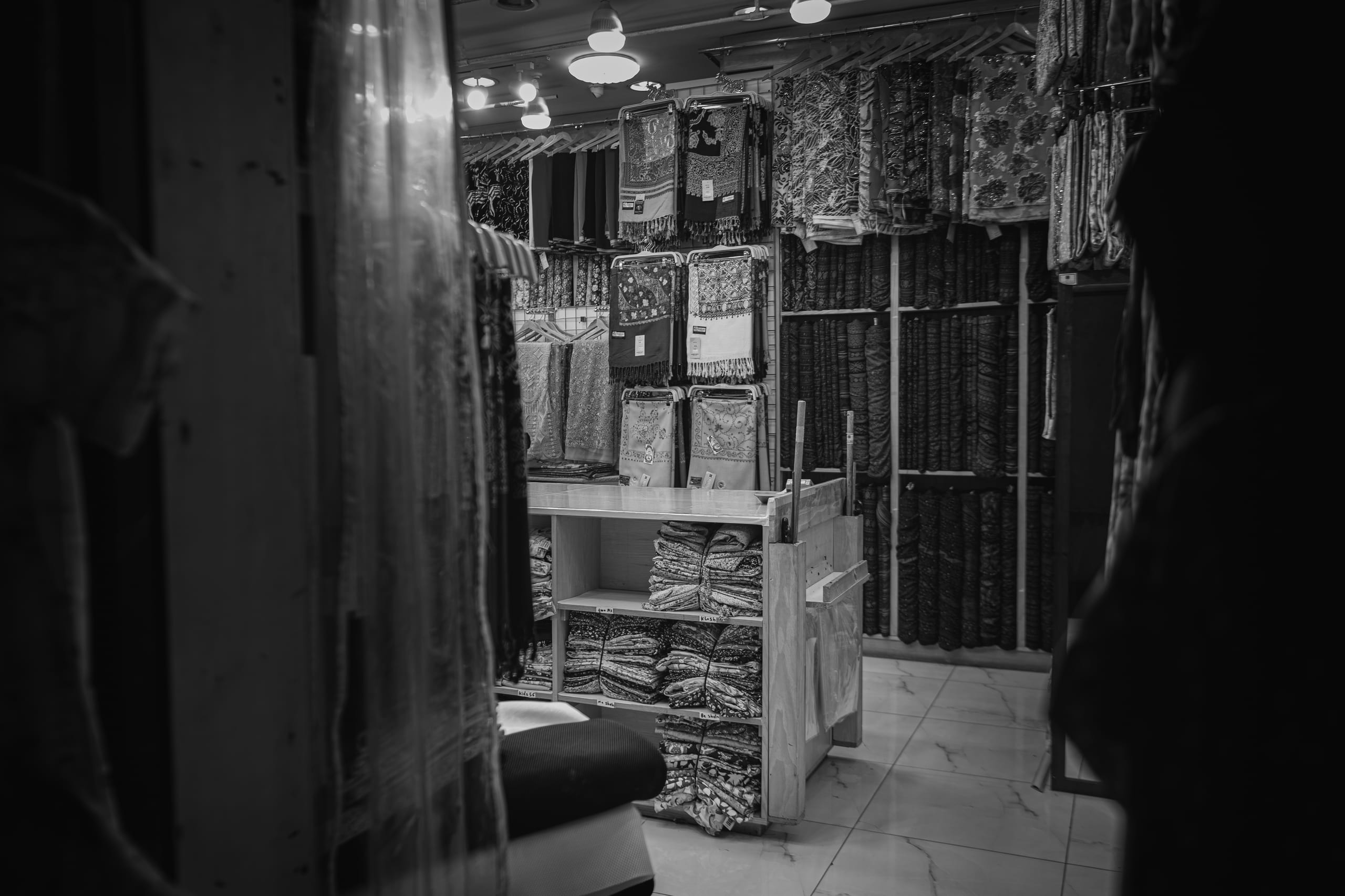 Monochrome interior view of a textile shop, featuring neatly folded stacks of fabric on wooden shelving and numerous hanging scarves under bright overhead lights, framed by dark silhouettes in the foreground.