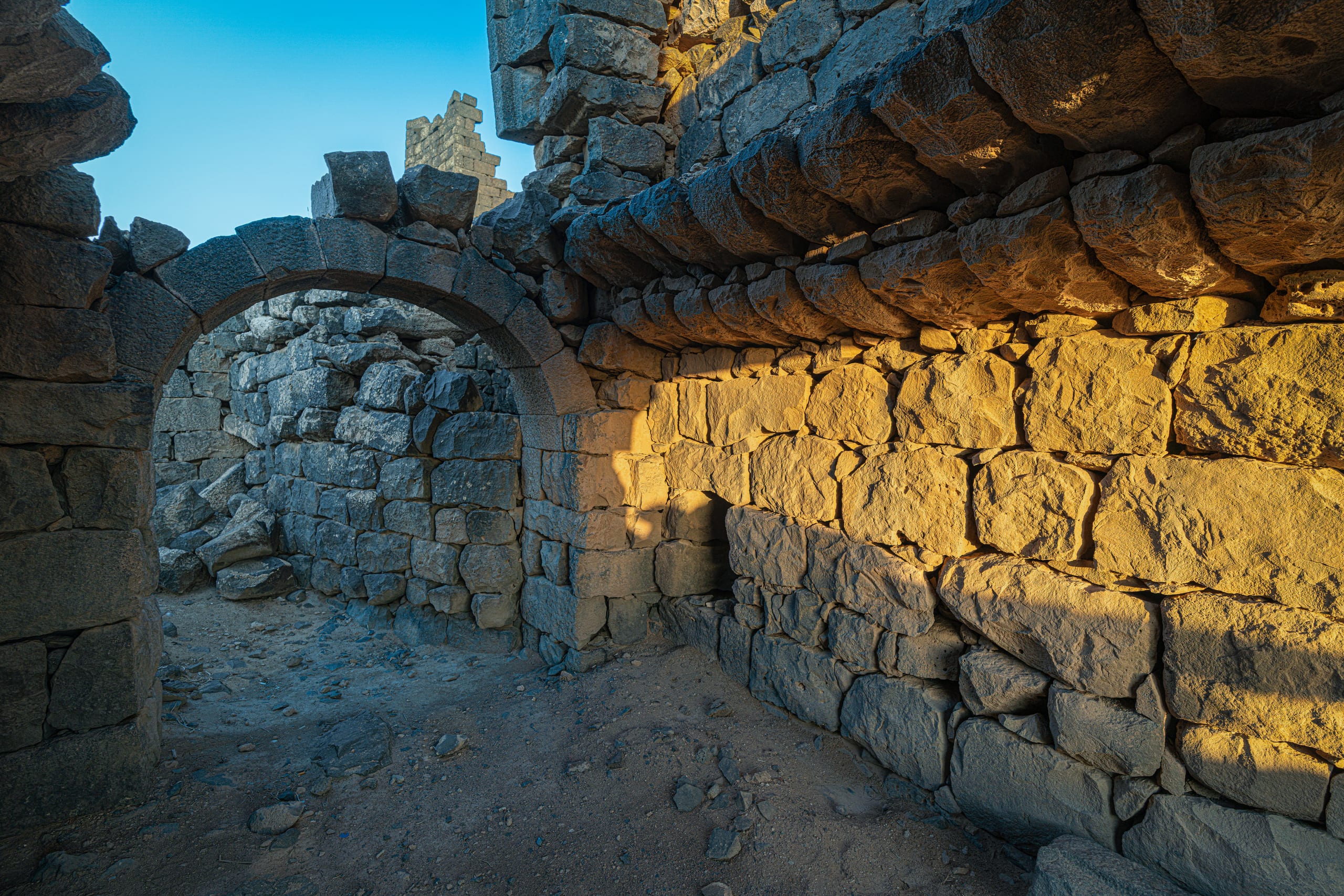 Interior view of ancient stacked stone walls and an arched entryway illuminated by golden sunset light casting strong shadows.