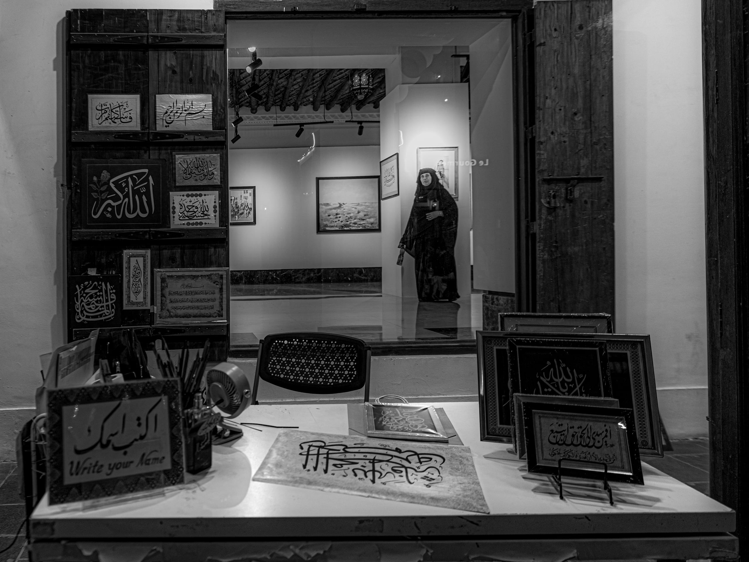 Monochrome interior photograph showing a framed view through a heavy wooden doorway into a gallery. A desk in the foreground holds framed Arabic calligraphy works, art supplies, and a sign in English and Arabic. In the background gallery, framed paintings hang on the walls and a figure stands in profile.
