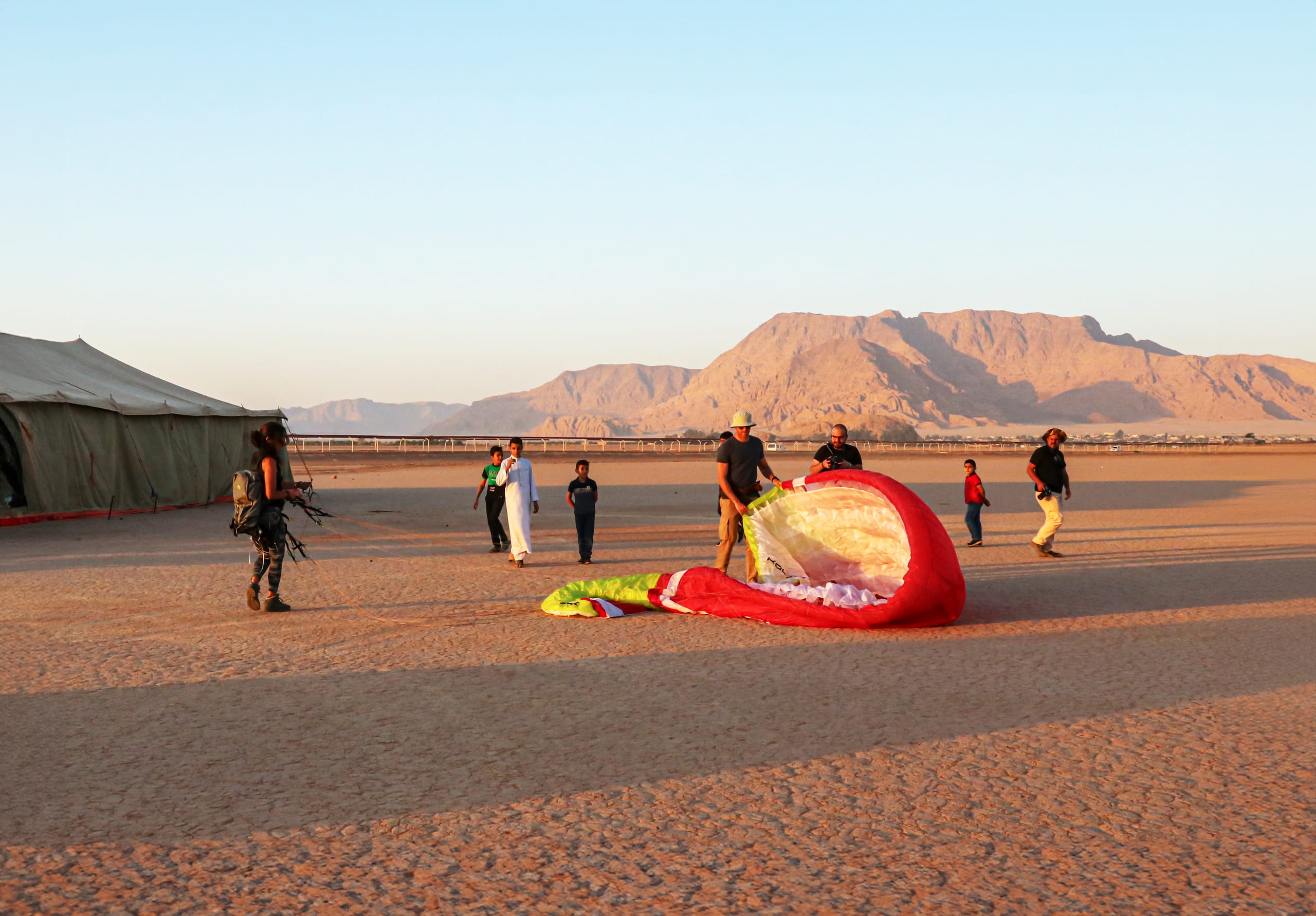 Wide shot of several adults and children gathered around a large red and green paraglider wing resting on sandy ground next to a large tent, with arid mountains in the background under clear daylight.