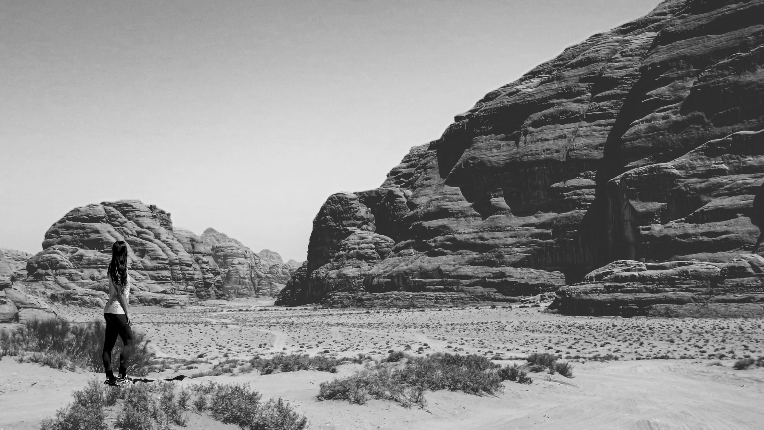 Black and white photograph showing a person standing on a sandy rise looking out toward massive rock cliffs across a desert canyon.