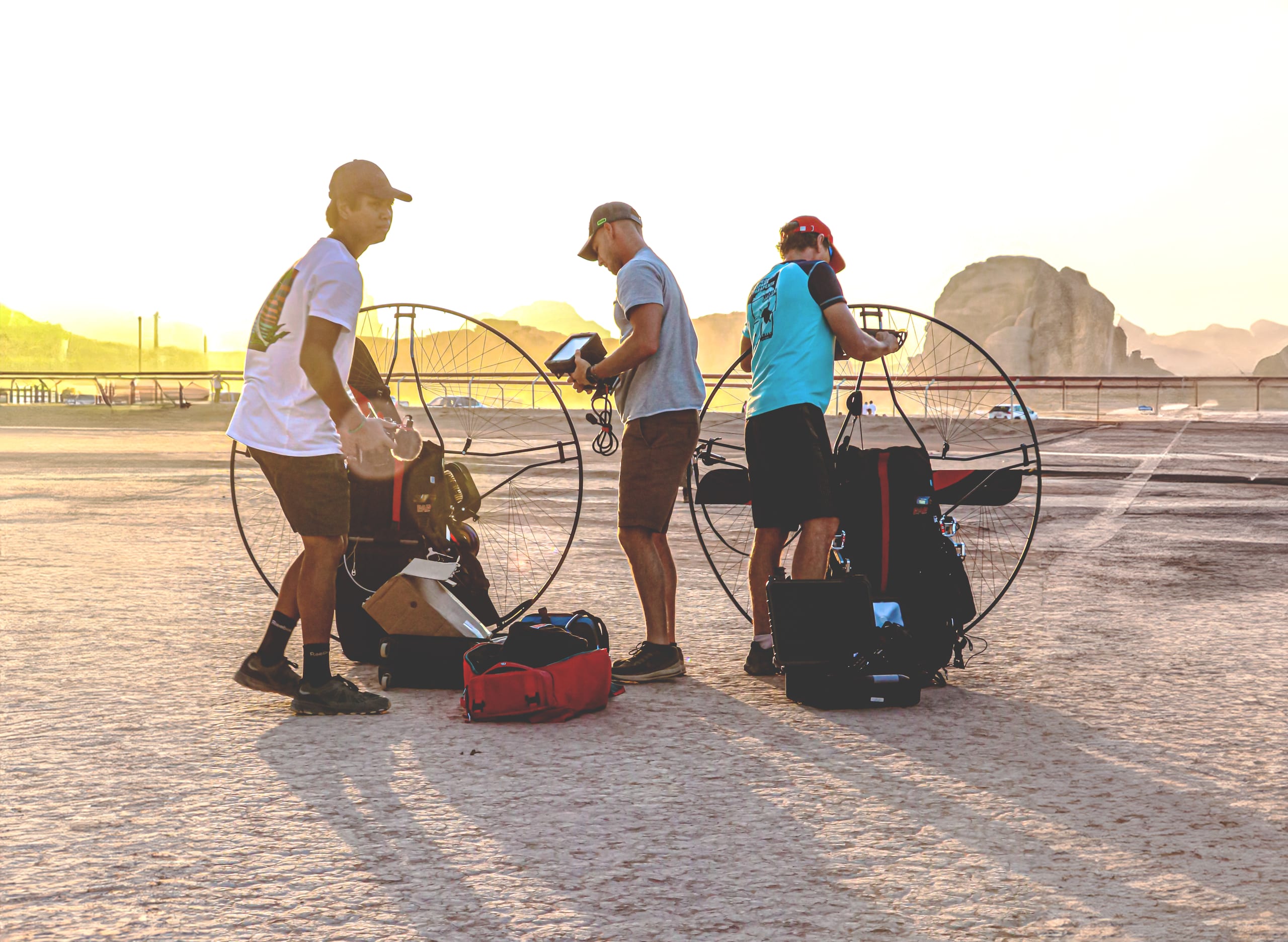 Three people stand outdoors on an open dusty plain in bright backlight, tending to large circular paramotor frames and gear packs on the ground.