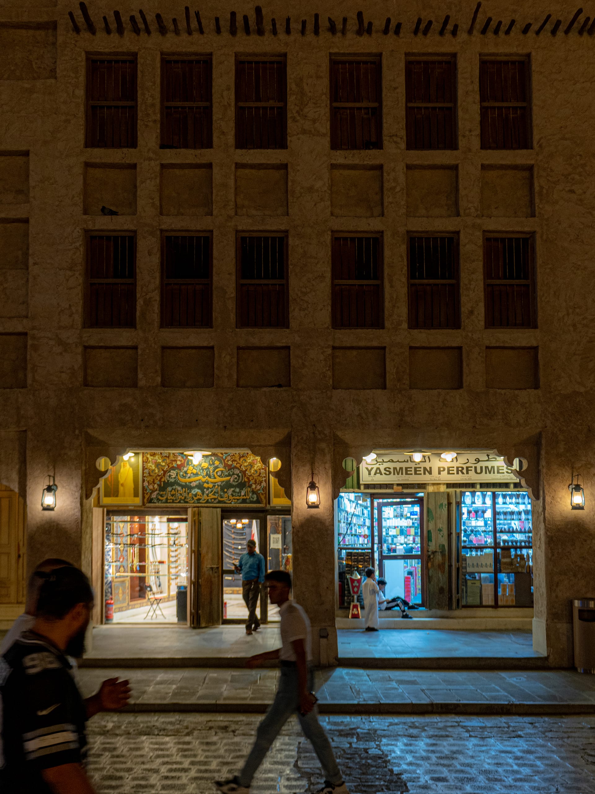 Night street scene showing pedestrians walking in front of a historic multi-story building with symmetrical wooden shutter windows and glowing storefronts at street level.