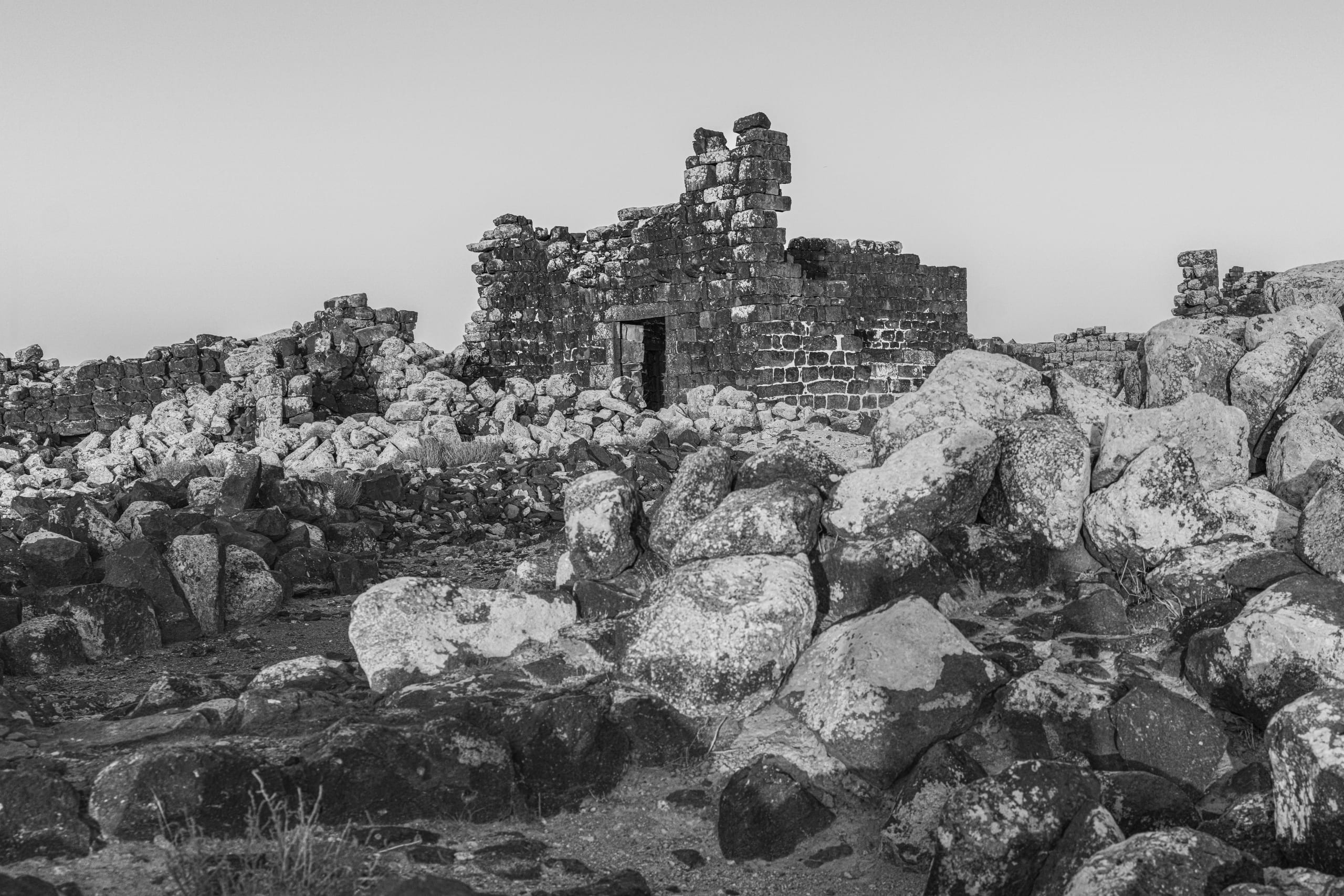 Black and white photograph of weathered dark stone ruins and fallen boulders in the foreground and midground, set against a pale uniform sky.