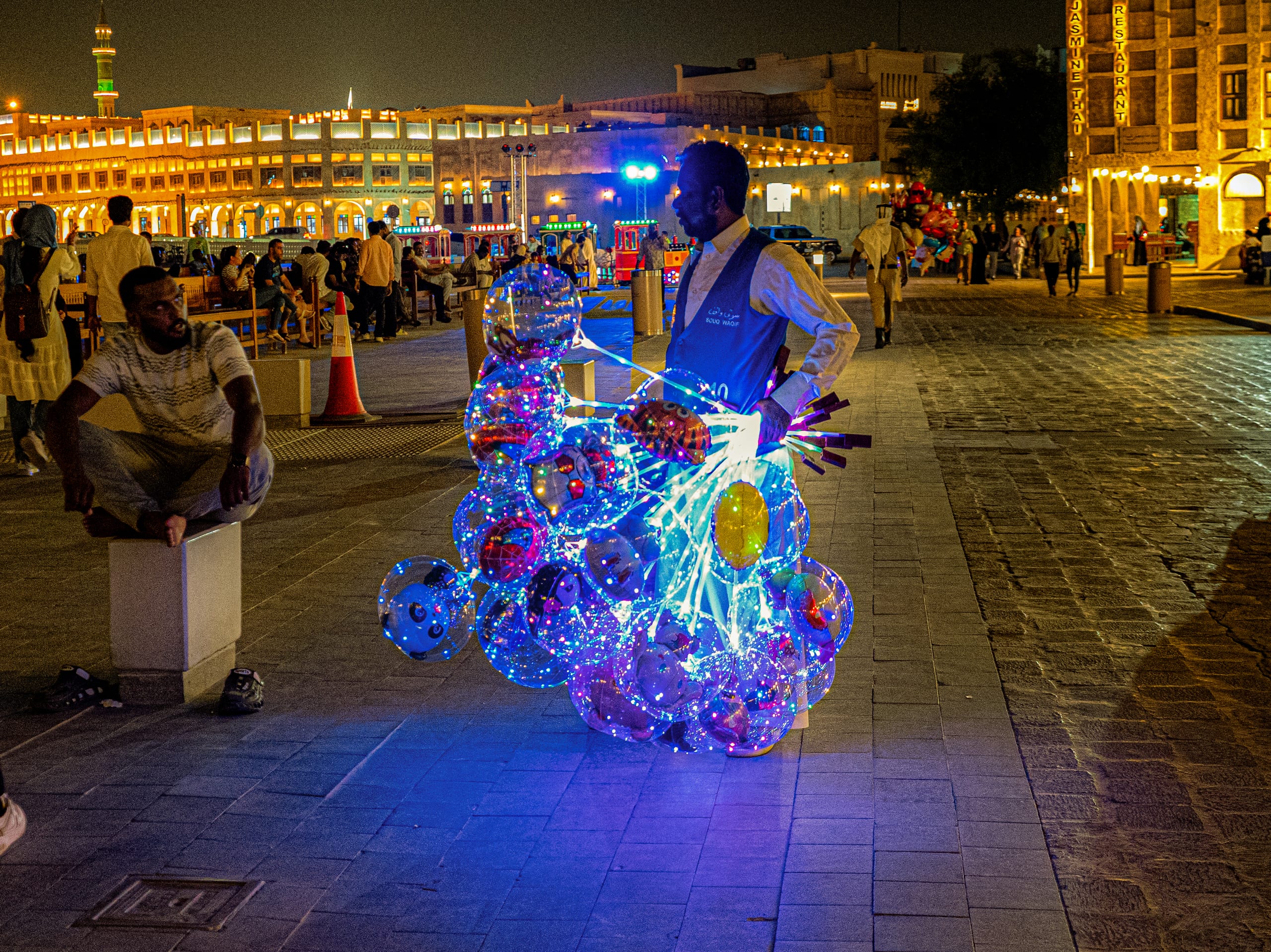 A vendor stands on a stone-paved plaza at night, holding a massive bunch of colorful, internally lit transparent balloons that cast a bright blue glow on the ground, while another man sits watching on a low block to the left.