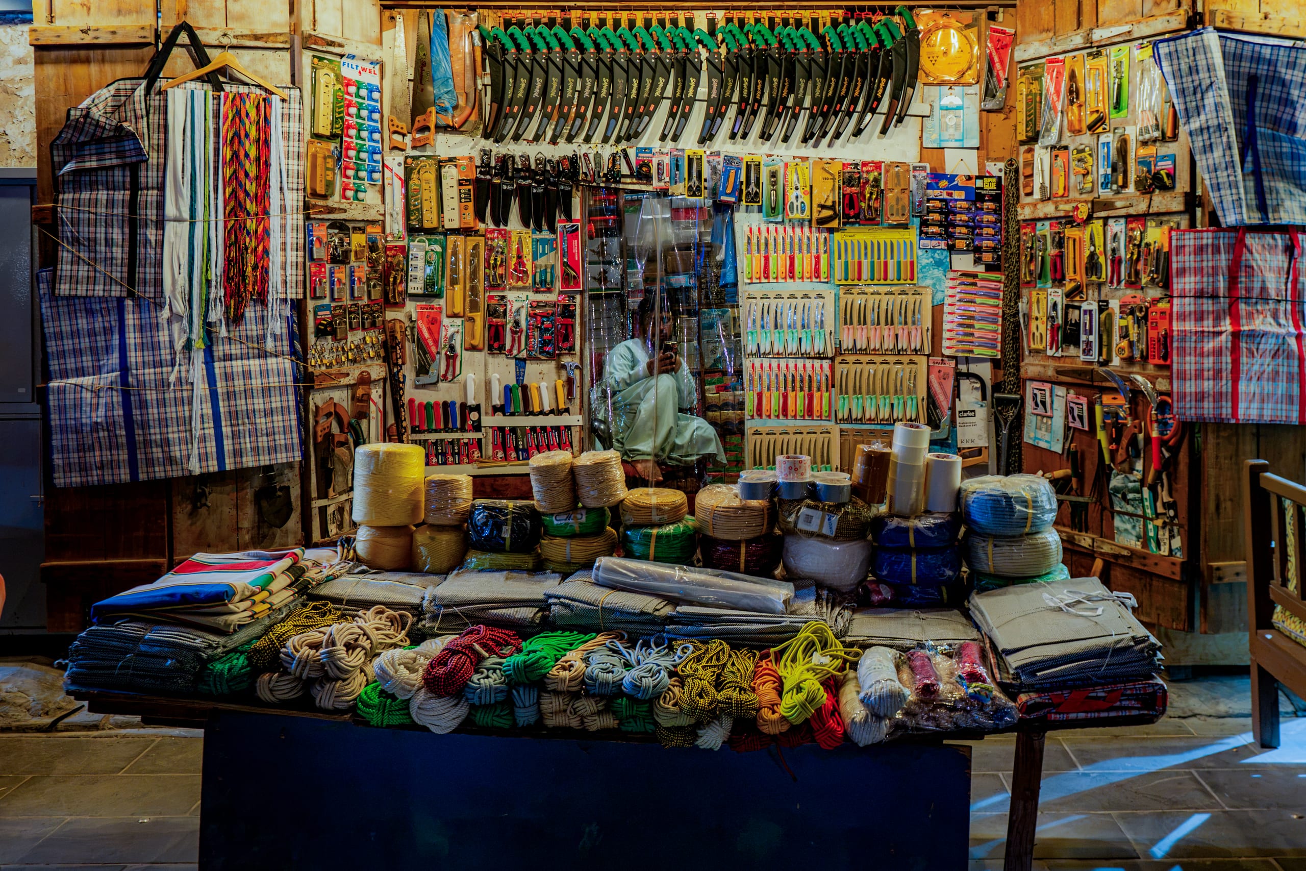 Interior view of a small shop featuring wooden walls completely covered in hanging knives, saws, and tools, with spools of rope and textiles neatly stacked on a wooden counter in the foreground and a person seated in the background.