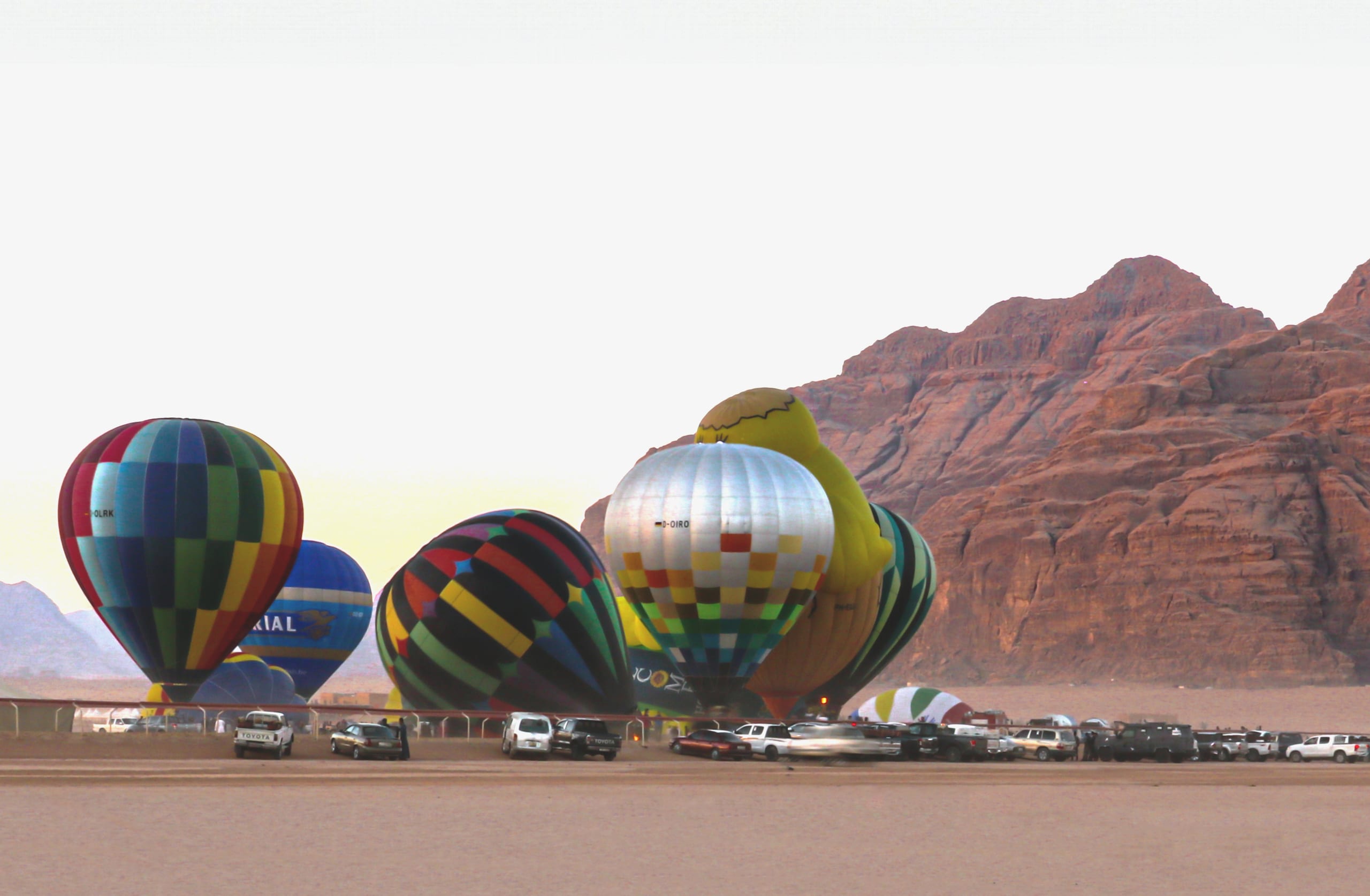 Several large hot air balloons in vibrant patterns are grounded on a desert floor beneath rugged rust-colored mountains, with parked vehicles lined up in the foreground.