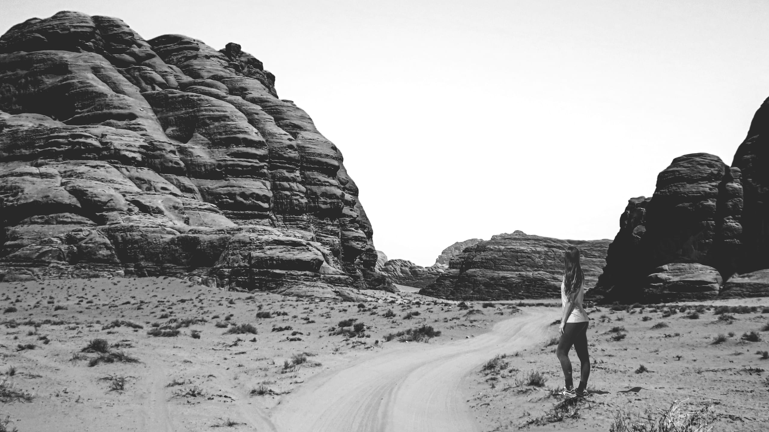 A woman stands on the right side of a wide, sandy desert path, looking toward towering dark rock formations under a bright white sky in a high-contrast monochrome photograph.