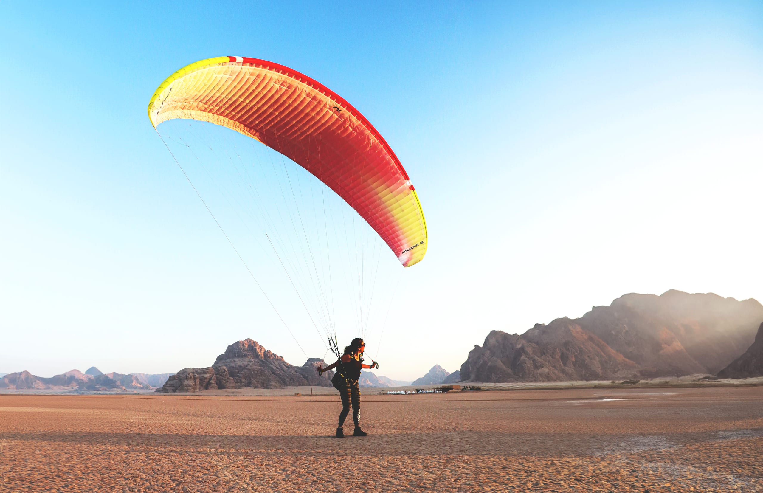 A paraglider holding a large red and yellow wing, standing on a flat desert floor with rocky mountains in the background under an open sky.