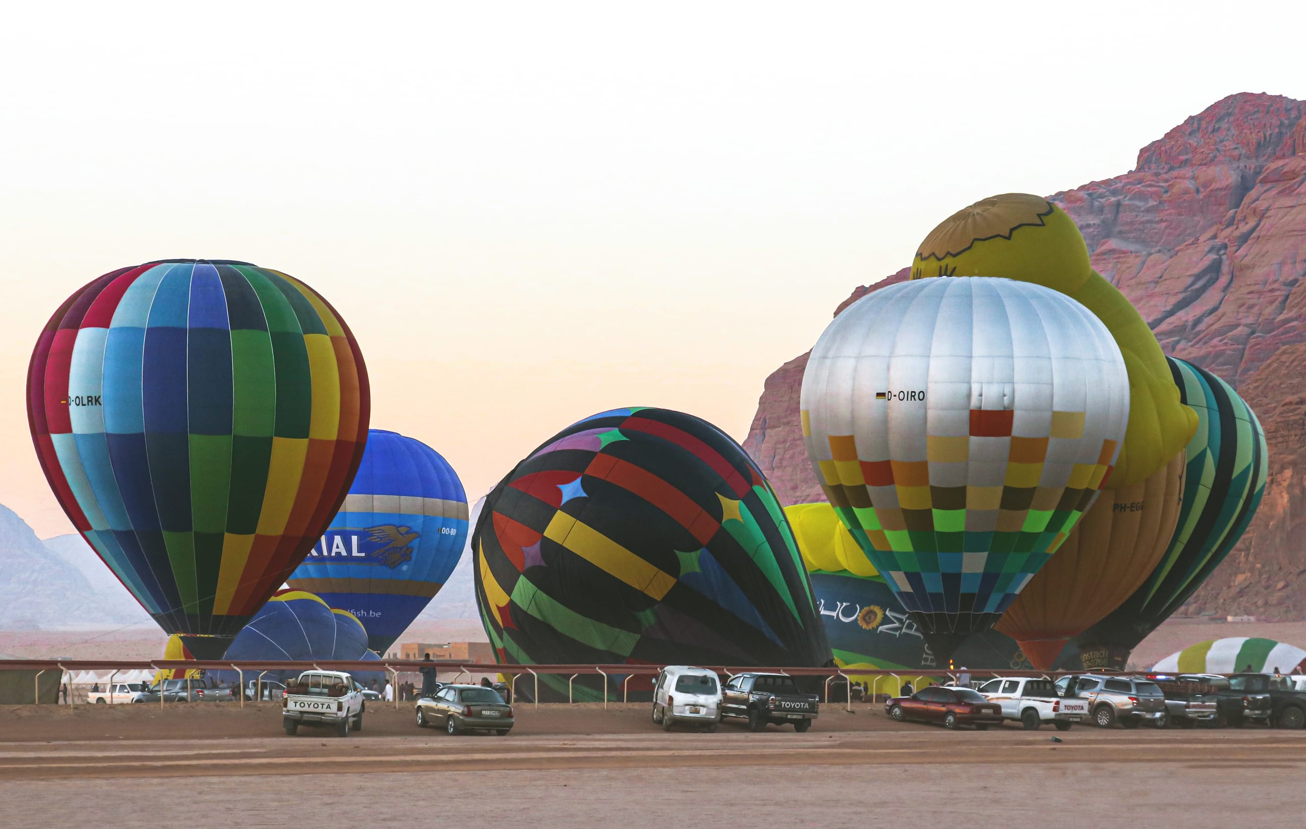 A line of colorful hot air balloons stands inflated on flat desert ground in front of rocky sandstone mountains under a clear sky, with parked pickup trucks visible in the foreground.