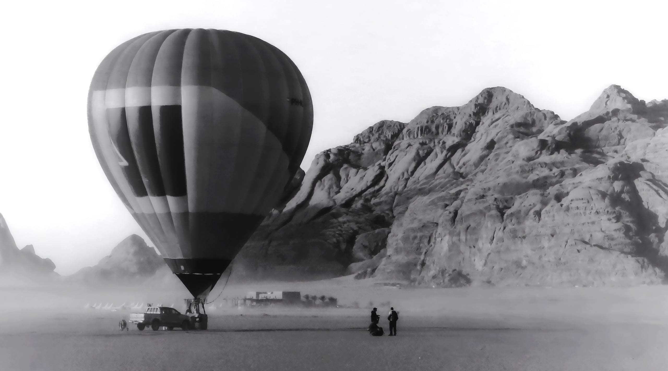 Monochrome photograph of a large hot air balloon grounded on a vast sandy plain, with two small figures standing nearby and towering rocky mountains in the background.